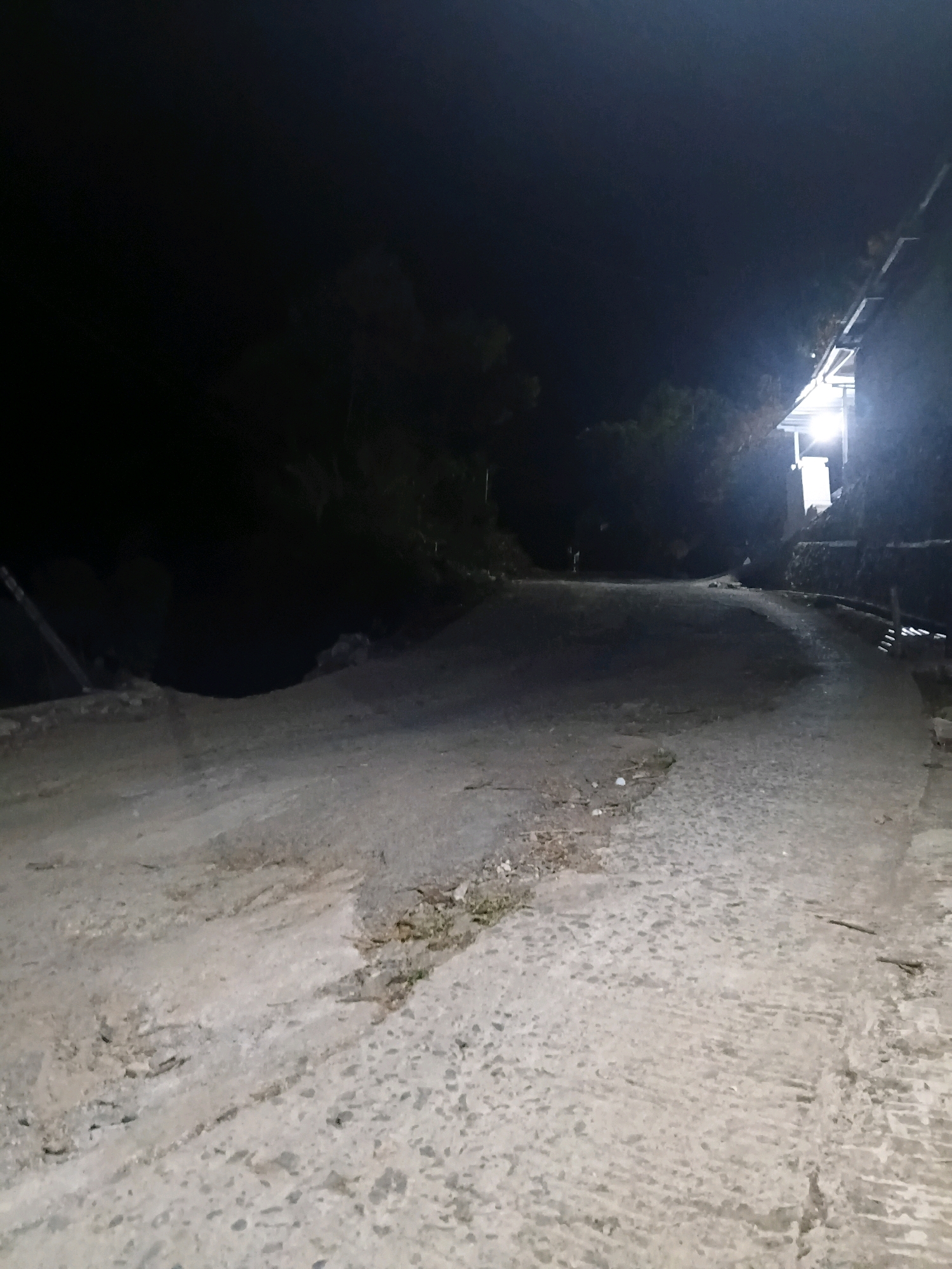A dimly lit, unpaved road winds uphill through a dark, wooded area. The road surface is a mix of dirt and rough, lighter-colored concrete or gravel. Debris, such as leaves and small stones, is scattered across the road. On the right side of the road, a building with a brightly lit window is visible, casting a glow onto the surrounding area. The building appears to be constructed with stone walls and a sloped roof. The overall scene suggests a rural or semi-rural setting in Sinduaji, Indonesia, at night. There are no people visible in the image.