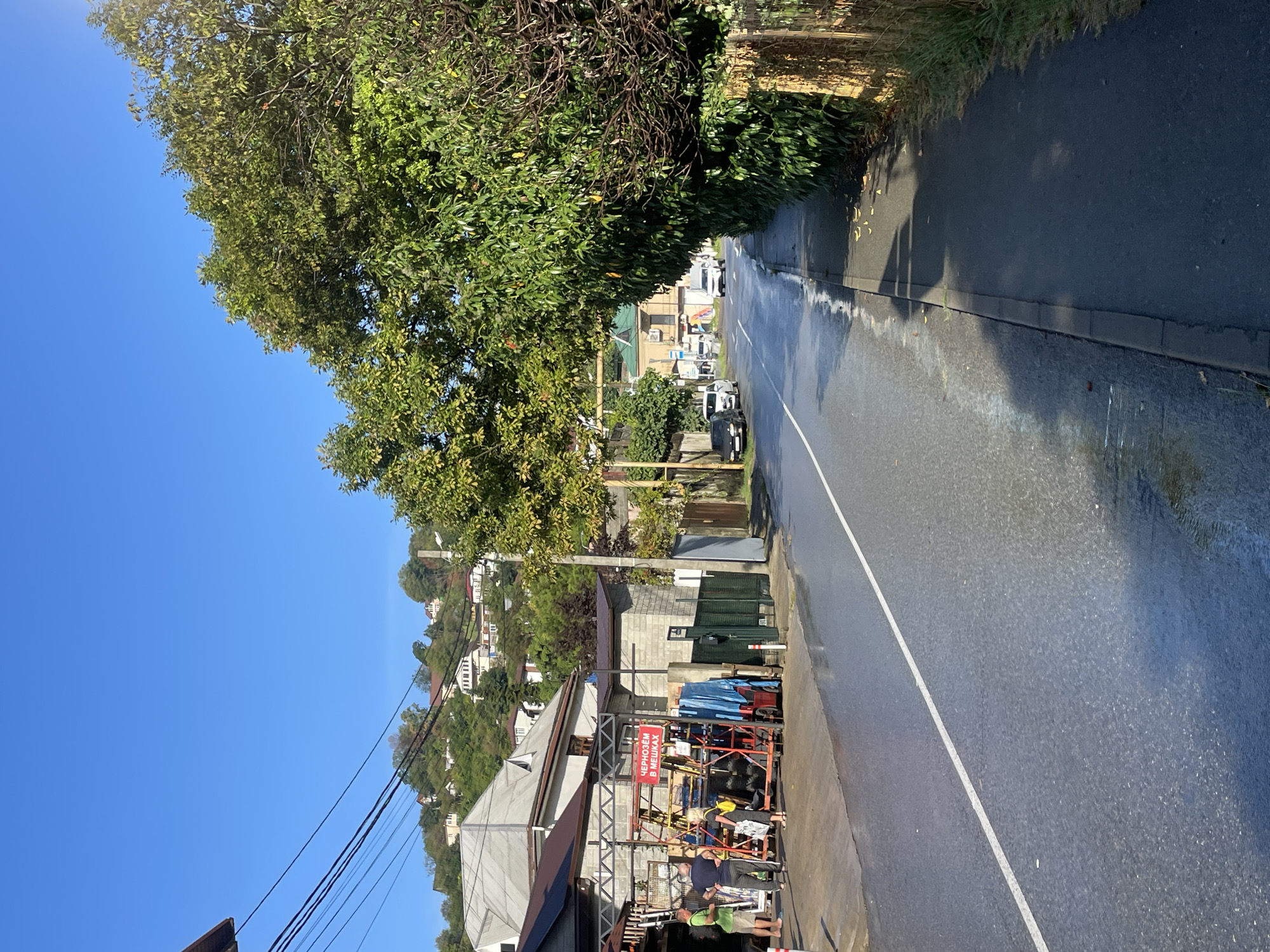 The image shows a street scene in Baranovka, Russia. The sky is clear and blue, indicating a sunny day. The road is wet, suggesting it may have recently rained or been cleaned. On the left side of the road, there is a building with a gray brick facade, and in front of it, there are several people standing around what appears to be a display of goods on wheeled racks. One rack has a red sign with white Cyrillic text that translates to "Chernozem in bags." There are also tarps and other materials visible.
Across the street, there is a sidewalk bordered by a fence and dense green foliage. Further down the road, there are several vehicles, including cars and a white van, driving on the wet asphalt. In the background, houses are visible on a tree-covered hillside, characteristic of the hilly terrain in Baranovka. The overall impression is of a commercial area along a road in a suburban or semi-rural setting.