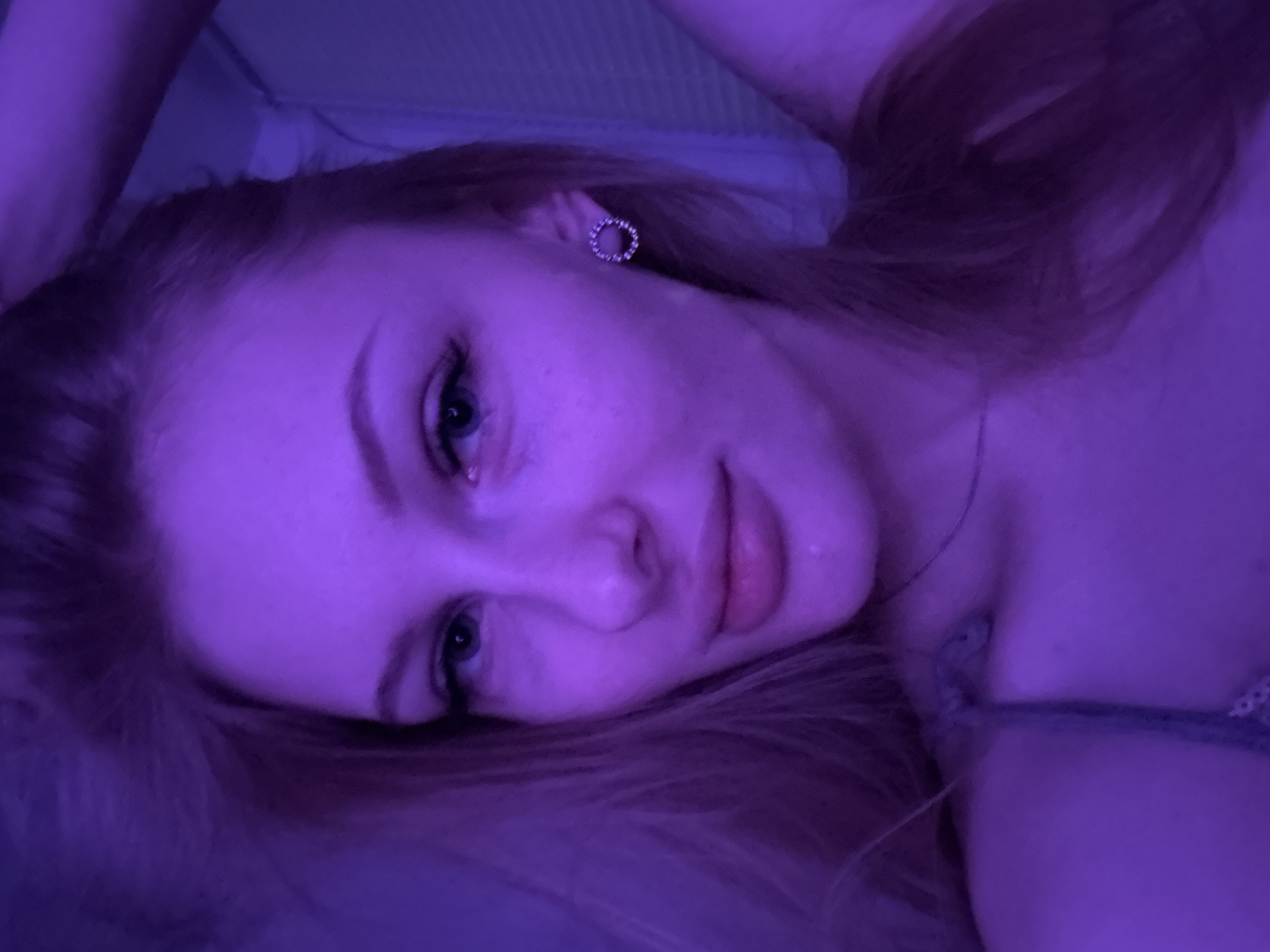A close-up portrait of a fair-skinned person with light brown hair under a purple light source. The person is lying down, angled towards the camera. A circular earring is visible on the upper left. A thin necklace rests against their neck. A small portion of a grey shirt is visible. No evidence suggests this photo was taken in Perm, Russia.