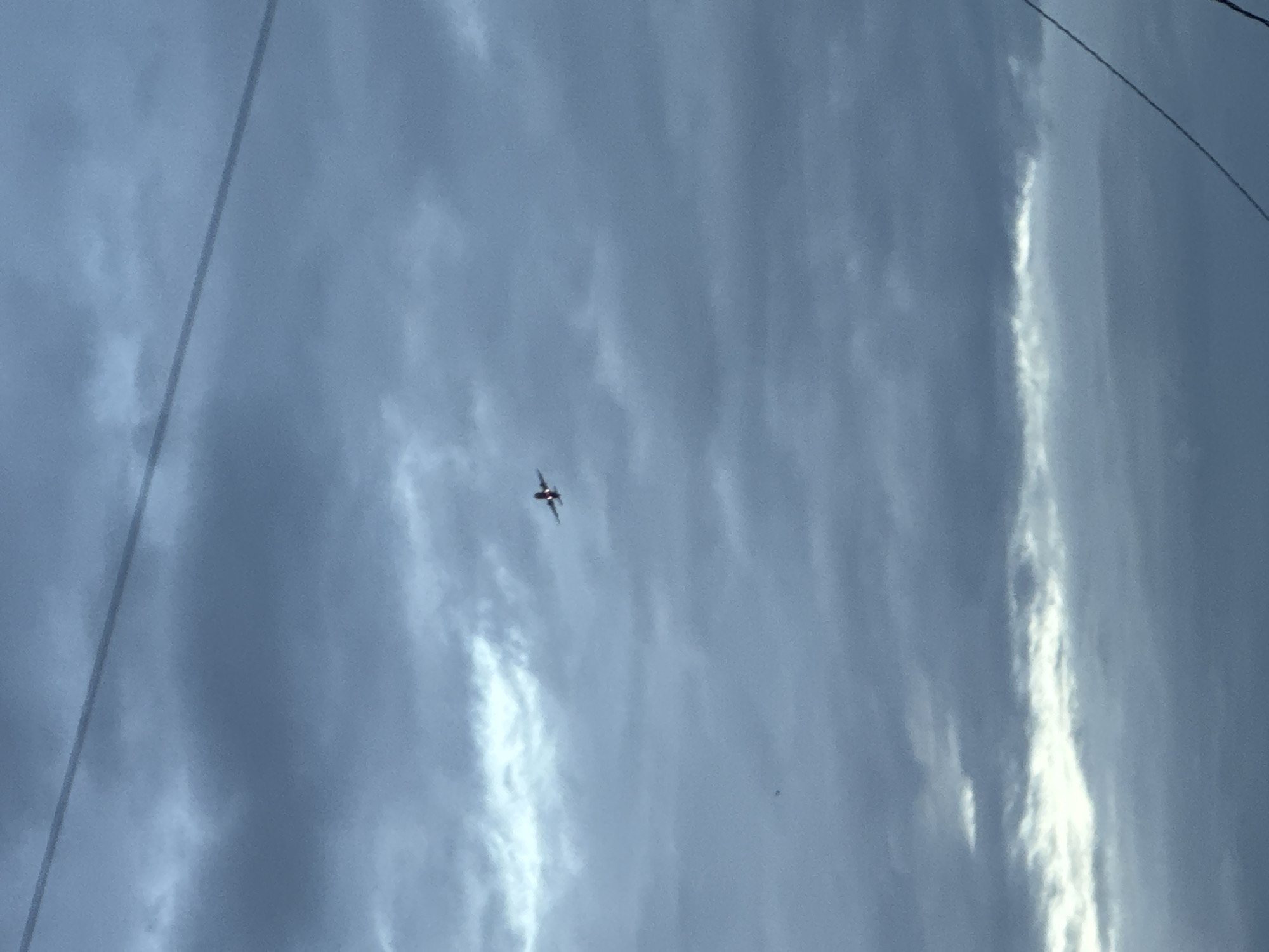 An airplane is seen flying high in a cloudy sky above Mikhaylovsk, Russia. The clouds are a mix of dark gray and lighter white, suggesting an overcast or stormy day. A thin, dark wire runs diagonally across the top portion of the image, and another wire is visible in the lower right corner. The airplane is small and centrally located in the frame, with its wings spread. The overall impression is a vast, cloudy expanse with a solitary aircraft navigating through it.
