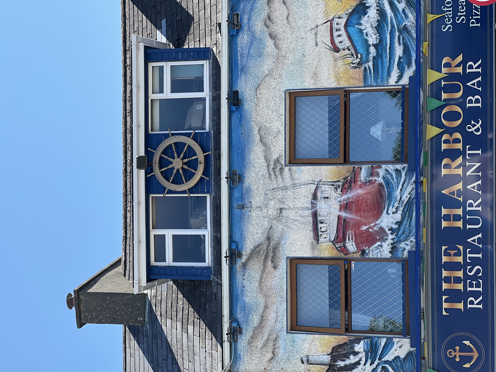 The image shows the facade of "The Harbour Restaurant & Bar" in Donegal, Ireland. The building features a mural painted on its exterior depicting ships at sea. The mural is vibrant, with blue waves and red and blue ships. Above the mural, there are two windows, and a ship's wheel is mounted between them. The roof is tiled, and a chimney is visible on the left side. Below the mural, a dark blue sign with gold lettering reads "THE HARBOUR RESTAURANT & BAR". Small colorful flags are strung across the top of the sign. The sky is clear blue.