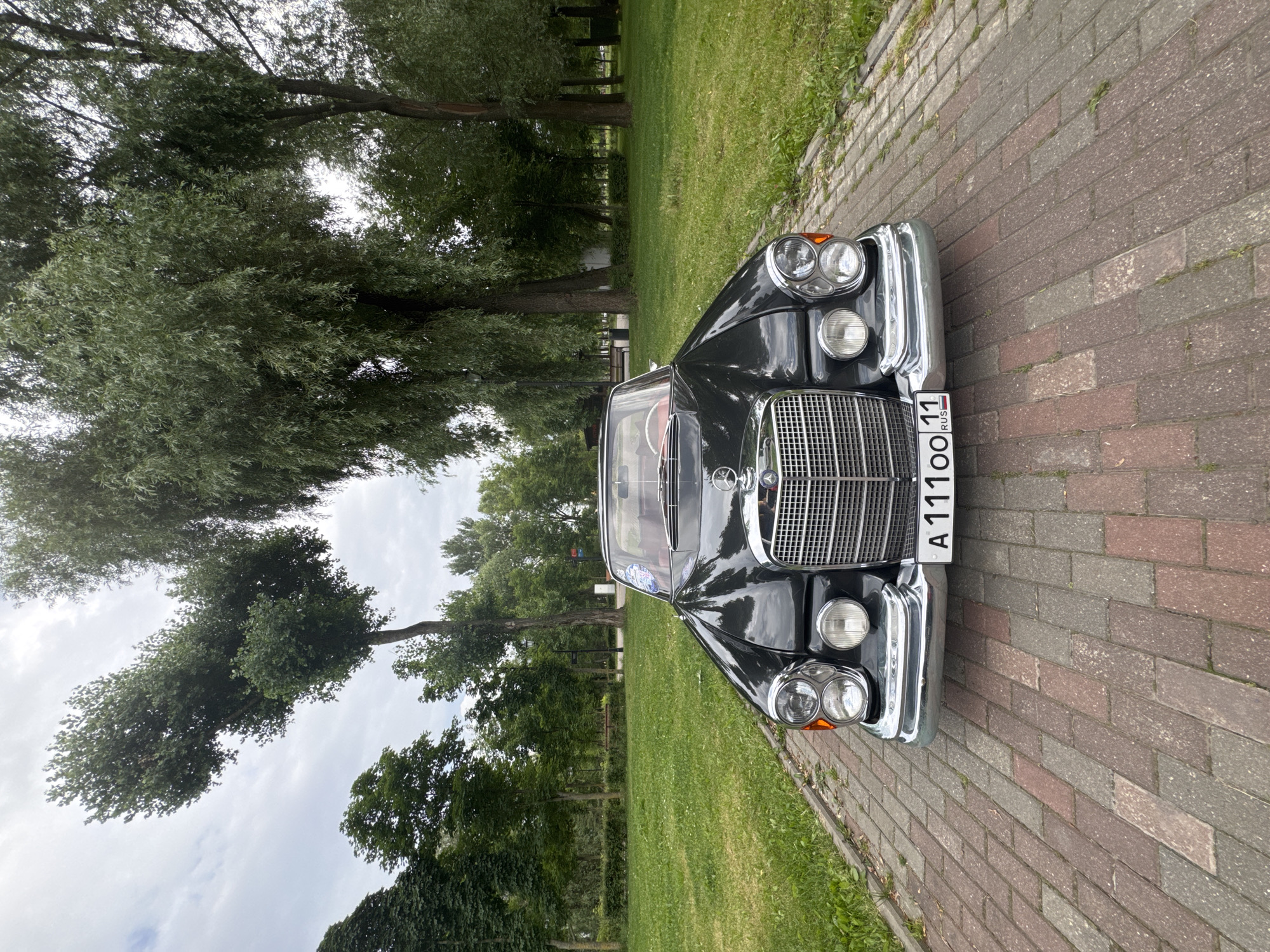 The image shows the front of a black Mercedes-Benz convertible parked on a brick pathway in a park-like setting. The car has a prominent chrome grille and headlights. The pathway is bordered by a well-maintained green lawn and mature trees. In the background, there are more trees, some with distinctive shapes, and a cloudy sky. The license plate of the car reads "A 111 OO 11 RUS". The setting appears to be a public park with benches visible in the distance, consistent with a location in Moscow, Russia. There are no people visible in the image.