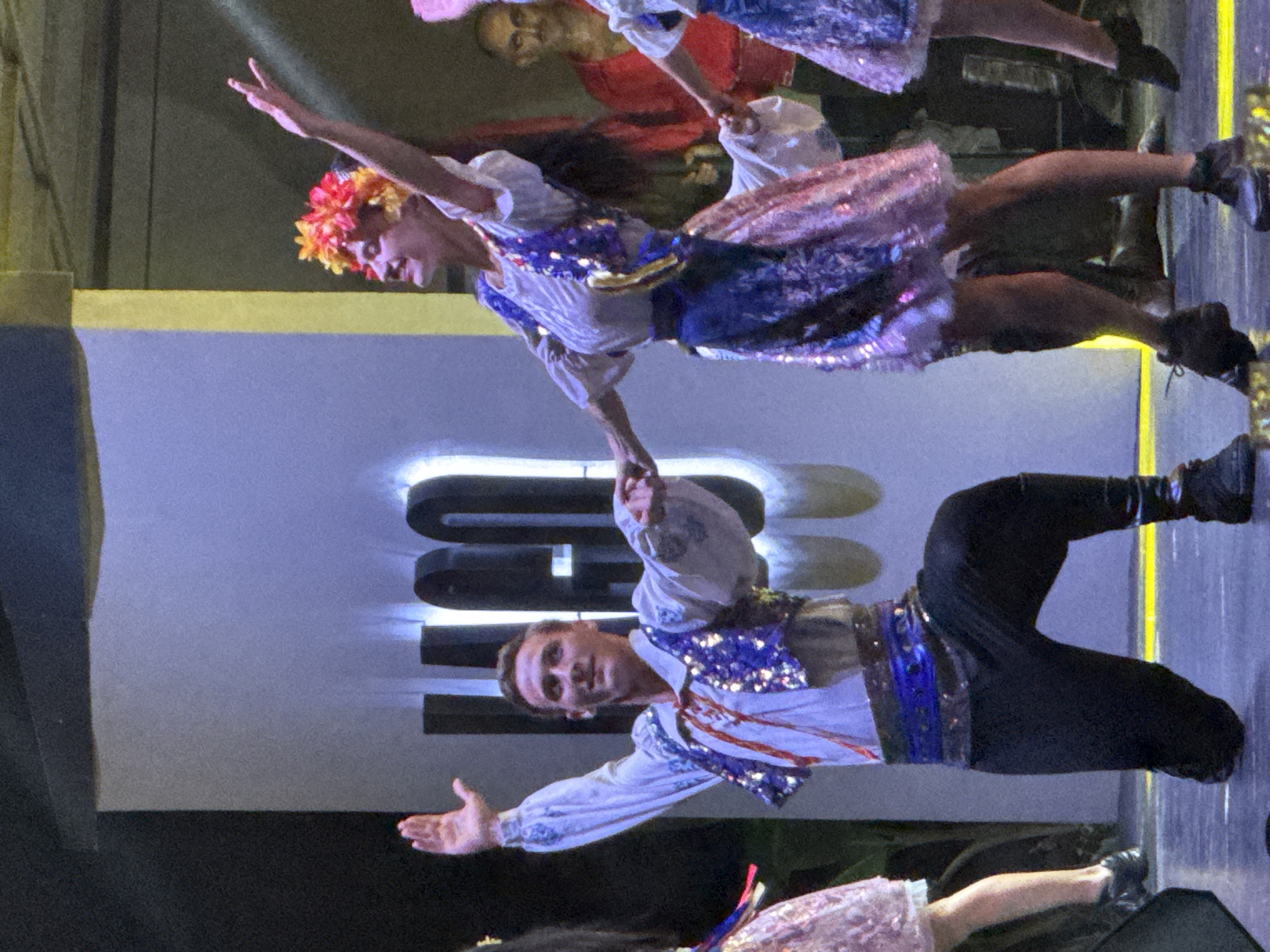 This is a medium shot of a performance on a stage, likely in Manavgat, Turkey, given the context. Two dancers are in the foreground, a man and a woman, both dressed in traditional, ornate costumes. The man, on the left, is kneeling on his right leg with his left leg extended. He wears a white shirt with embroidered details, a sequined blue vest adorned with red trim, black pants, and dark boots. His right arm is raised, and his left arm is extended to his side. The woman, on the right, is in mid-dance pose with her left arm raised and her right arm extended towards the man. She wears a white top, a sequined blue vest over it, a colorful skirt, and a floral headdress. In the background, other performers are visible, including a man in a red shirt and another in a blue and white outfit, suggesting a group performance. The stage backdrop features large, stylized black letters that appear to spell "MANAVGAT," illuminated by soft blue and white lighting. Yellow stage lights are visible on the floor, and the overall environment suggests an outdoor or indoor venue set up for a cultural or entertainment event.