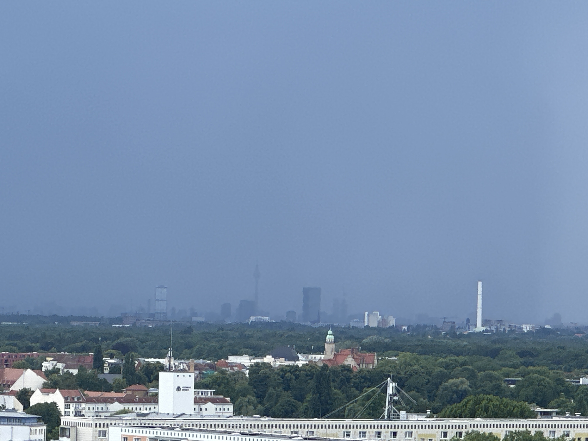 The image shows a hazy cityscape of Berlin, Germany, from a high vantage point. In the foreground, there are several buildings with red-tiled roofs and white facades. A large white building with a logo is prominent in the center-right. Beyond these buildings, a dense green forest stretches across the landscape. In the distance, the silhouette of the Berlin skyline is visible through the haze, featuring various skyscrapers and structures, including what appears to be the Fernsehturm (TV Tower) and a tall, white smokestack. The sky is a uniform, overcast blue. No people are visible in the image.