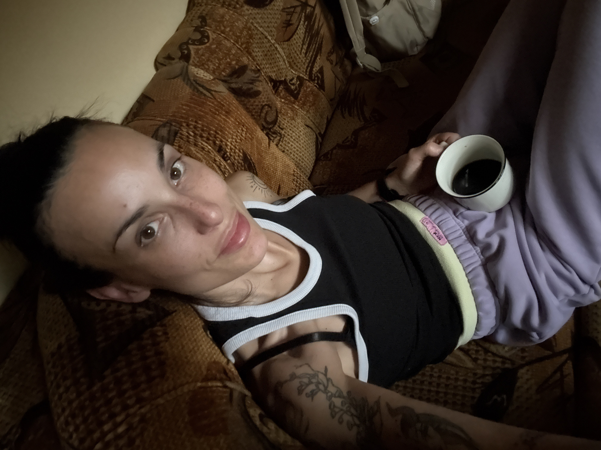 A woman is reclining on a brown patterned sofa with her head near the left edge of the frame and her legs towards the right. She is wearing a black tank top with white trim and lavender sweatpants with a yellow and pink stripe along the waistband. In her right hand, she is holding a white mug filled with a dark liquid, possibly coffee. Her hair is pulled back, and she is looking towards the camera. Her left arm is visible with tattoos. The location appears to be indoors. The city of Pleven, Bulgaria cannot be determined from this image.