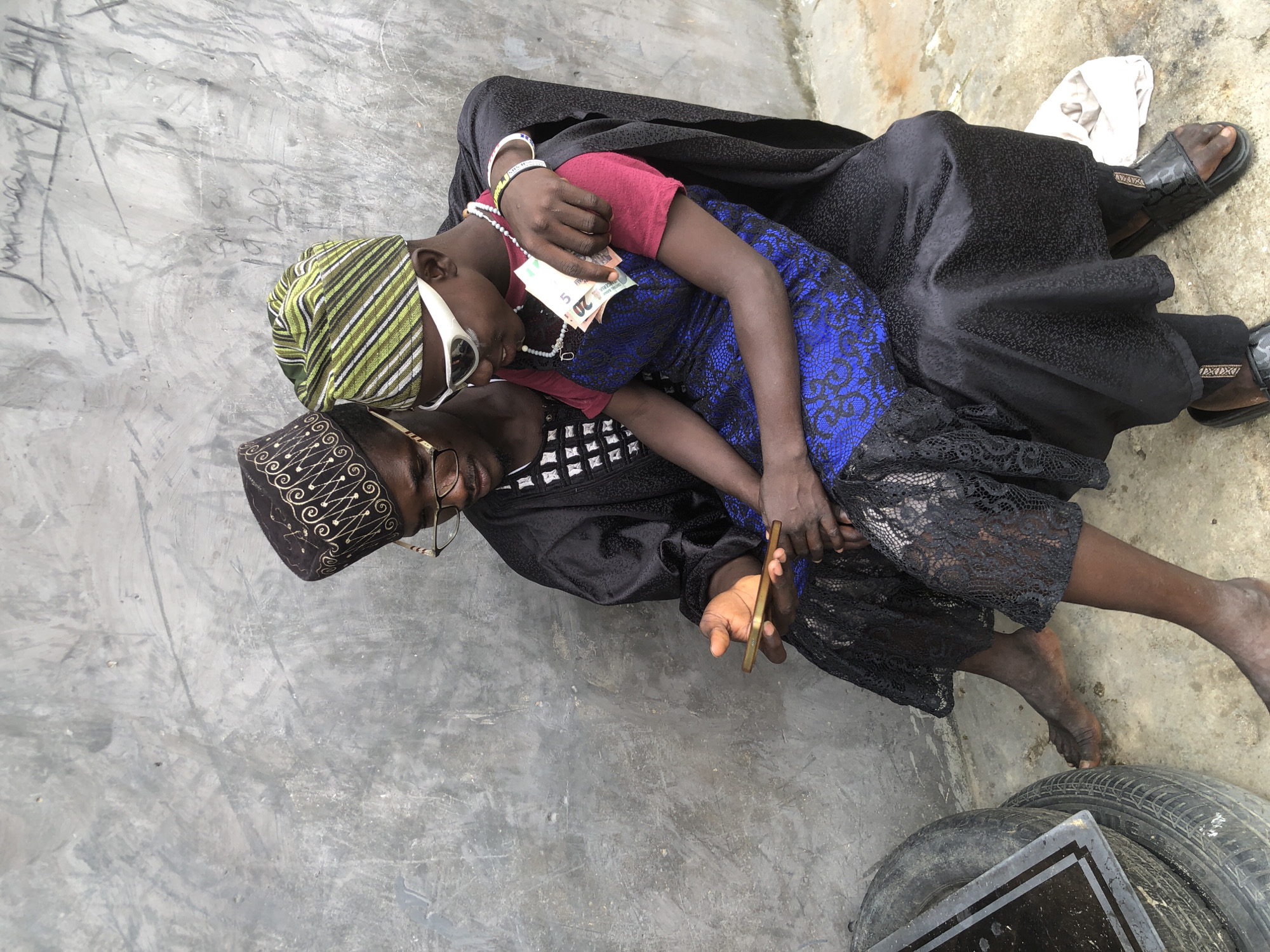 Two individuals, an adult male and a younger individual, are seated outdoors in Ado, Nigeria, against a textured gray concrete wall. The male, on the left, wears a black cap with gold embroidery, glasses, and a black robe with intricate white patterns on the chest. He holds a gold-colored smartphone in his right hand, seemingly showing it to the person next to him. The younger individual, to the right and nestled against the male, wears a green and white striped cap, white sunglasses, a red t-shirt, and a blue lace-patterned dress. They are holding a few Nigerian Naira banknotes, a 20 and a 5. The younger individual's left arm is around the male, who has his right arm around the younger person's shoulders. They are both barefoot, with the male wearing black slides and the younger individual also barefoot. In the foreground to the left, two stacked tires are visible. A white cloth is on the ground to the right. The lighting suggests it is daytime.
