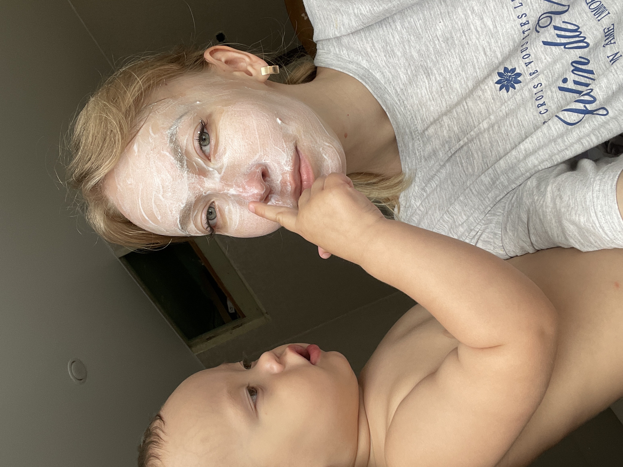 A selfie angled upward depicts a woman with blonde hair and a white facial mask on her face, partially obscured by a baby's hand touching her nose. The baby, with fair skin, is visible in the lower portion of the frame, lying down and looking up at the woman. The woman is wearing a gray heathered top with blue lettering and a flower design. The background includes a light gray wall and a dark green window. No specific details indicate the location is Perm, Russia.