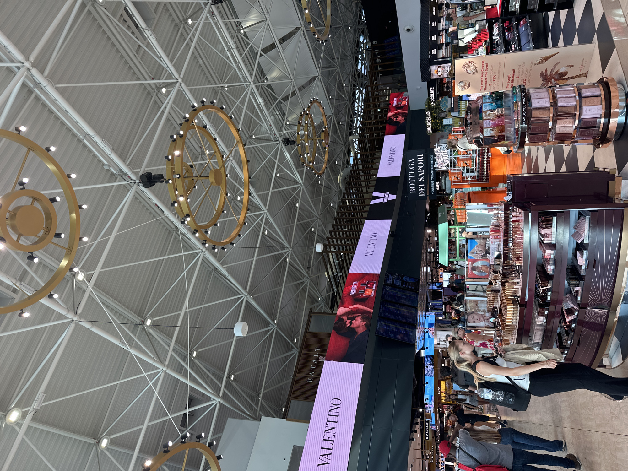 This image captures a bustling scene inside a duty-free shop at Fiumicino Airport in Fiumicino, Italy. The expansive ceiling structure with its intricate metal framework and large, circular gold chandeliers is a prominent feature of the environment. Large digital screens display advertisements for luxury brands such as Valentino and Bottega dei Sapori. In the foreground, a woman with blonde hair, wearing a white tank top and black pants, is intently examining a display of makeup and perfume products. She has a black backpack on and is holding a phone in her hand. Several other individuals are visible in the background, browsing the various retail displays and navigating the airport concourse. The floor is a distinctive black and white checkered pattern. The overall impression is one of a modern, well-lit, and commercially active airport retail area.