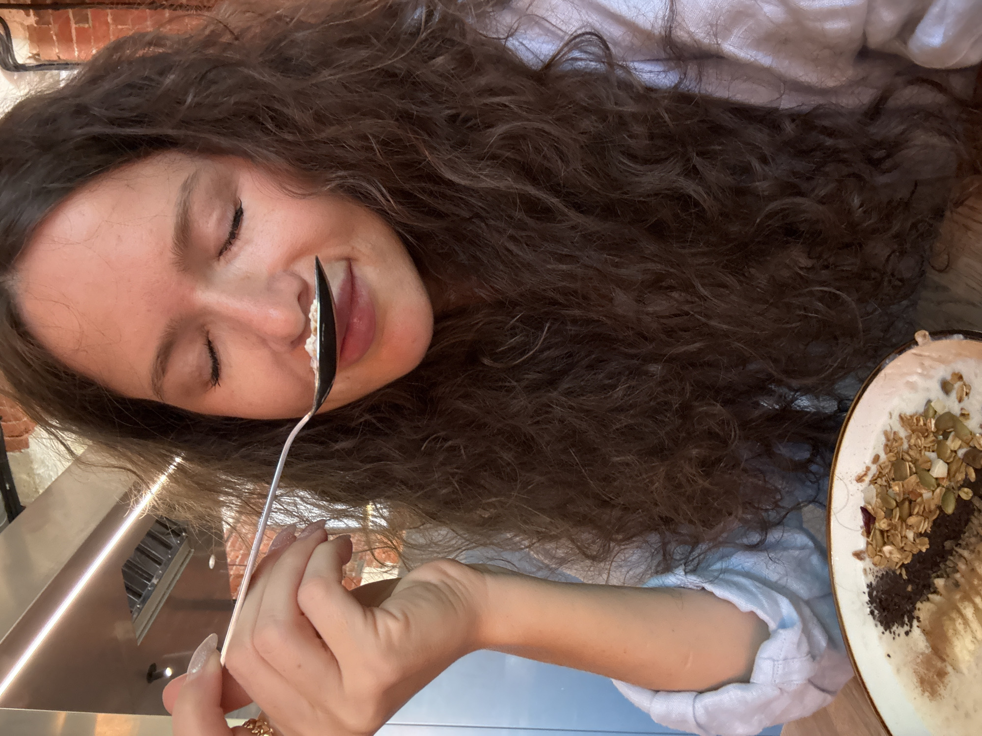In a close-up shot, a woman with long, curly brown hair is captured as she brings a spoonful of food to her mouth. Her eyes are closed, and her facial expression suggests she is savoring the taste. She is wearing a light blue garment. In the foreground, a bowl of oatmeal is visible, topped with granola, seeds, and what appears to be cocoa powder. The background is blurred, but hints of industrial elements, possibly ventilation systems, and brickwork suggest an urban, perhaps loft-style setting. The scene is likely in Moscow, Russia, given the context.