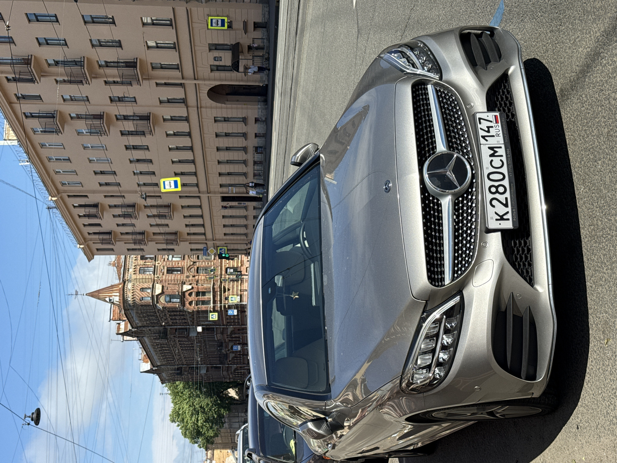 In Saint Petersburg, Russia, on a sunny day, a silver Mercedes-Benz C-Class sedan is parked on the side of a city street. The car, with license plate K280CM 147, is facing the viewer, showcasing its front grille with the Mercedes-Benz logo and its headlights. In the background, an old brick building with ornate architecture stands to the left, and a multi-story apartment building with a beige facade is on the right. Several yellow road signs with pedestrian icons are visible, and a traffic light shows green. A few pedestrians are walking on the sidewalk in the distance. The sky is blue with scattered clouds, and power lines are strung across the frame.