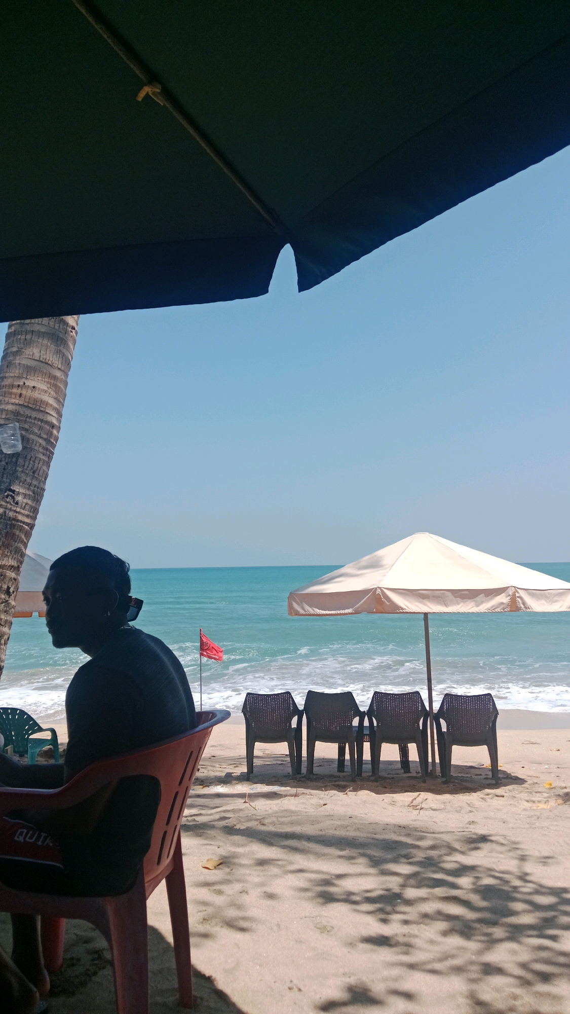 A silhouette of a person is visible in the foreground, seated in a red plastic chair on a sandy beach. The person is facing left, looking out at the ocean. Behind the person, a large dark umbrella covers part of the scene. A palm tree is to the left, its trunk partially visible. In the middle ground, several dark plastic chairs are arranged on the sand facing the ocean. A small red flag on a pole stands near the water's edge. A beige beach umbrella is open, providing shade for the chairs. The ocean is a vibrant turquoise with gentle waves breaking on the shore. The sky is clear and blue. The scene appears to be a relaxed beach setting in Kuta, Indonesia, likely during daytime.