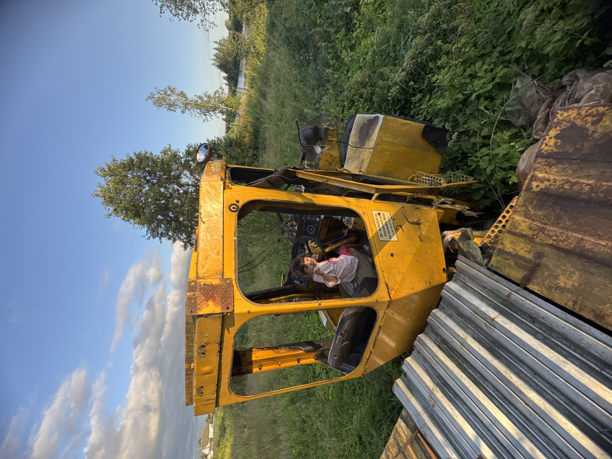 A young girl is sitting in the driver's seat of a yellow, weathered tractor cab, which is surrounded by tall green grass and vegetation. She is wearing a pink shirt and red pants. The cab itself appears to be part of a derelict piece of machinery, with visible rust and disuse. In the background, there are trees and a body of water under a blue sky with scattered clouds. The overall scene suggests a rural or abandoned setting. This location is identified as Innolovo, Russia.
