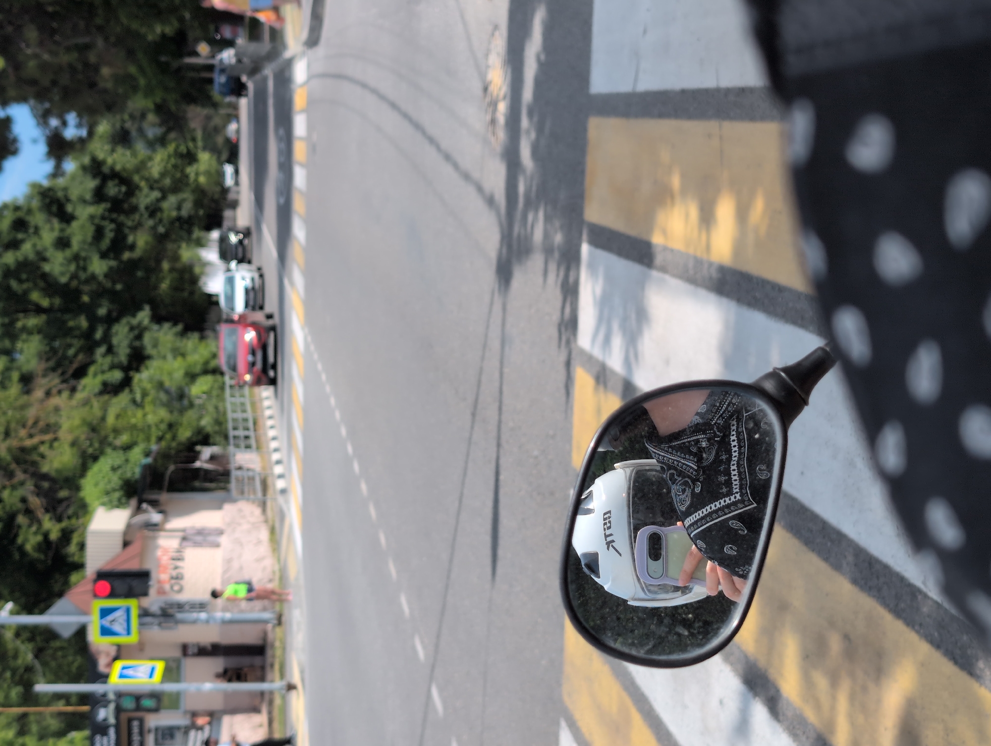 In Divnomorskoye, Russia, a reflection in a motorcycle's side mirror shows a person wearing a white helmet and a black bandana print shirt, holding a light purple mobile phone. The person is reflected against a backdrop of a street scene, with cars on a road, trees lining the street, and a pedestrian crossing. A traffic light is visible on the left side of the image, showing red. The reflection also captures part of the motorcycle's handlebar and the rider's patterned clothing. The environment appears to be a sunny day in a town or city.