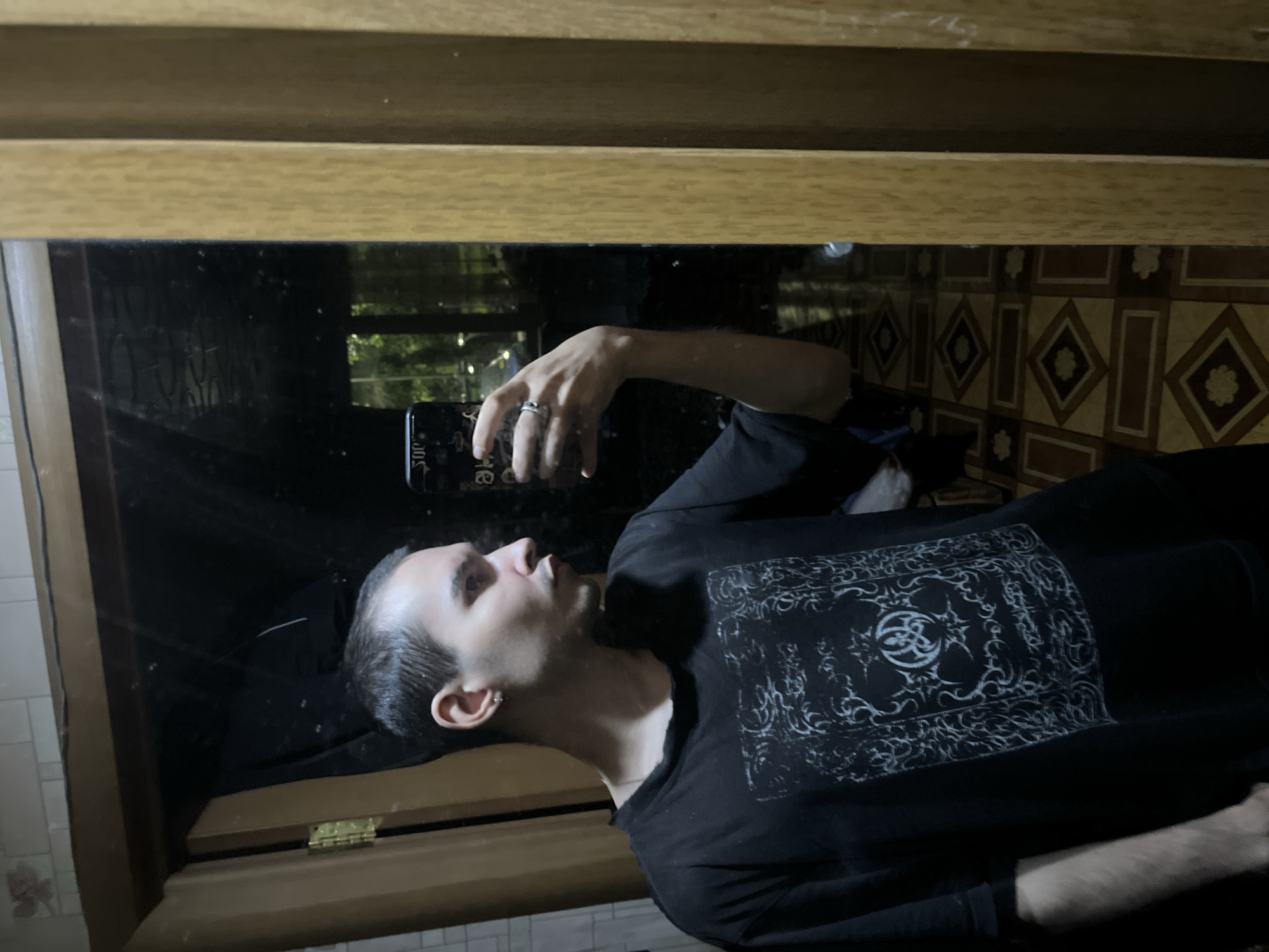 A young man with short hair and a hoop earring in his left ear is taking a selfie in a mirror. He is wearing a black t-shirt with a white ornate design on the front. His right hand is holding a smartphone, and a ring is visible on his ring finger. The background shows a tiled floor with a diamond pattern and a doorway with a wooden frame. The overall impression is that the photo was taken indoors, possibly in a bathroom or hallway, given the tiles and the presence of a mirror. There is no specific information in the image to indicate the location is Kazan, Russia.