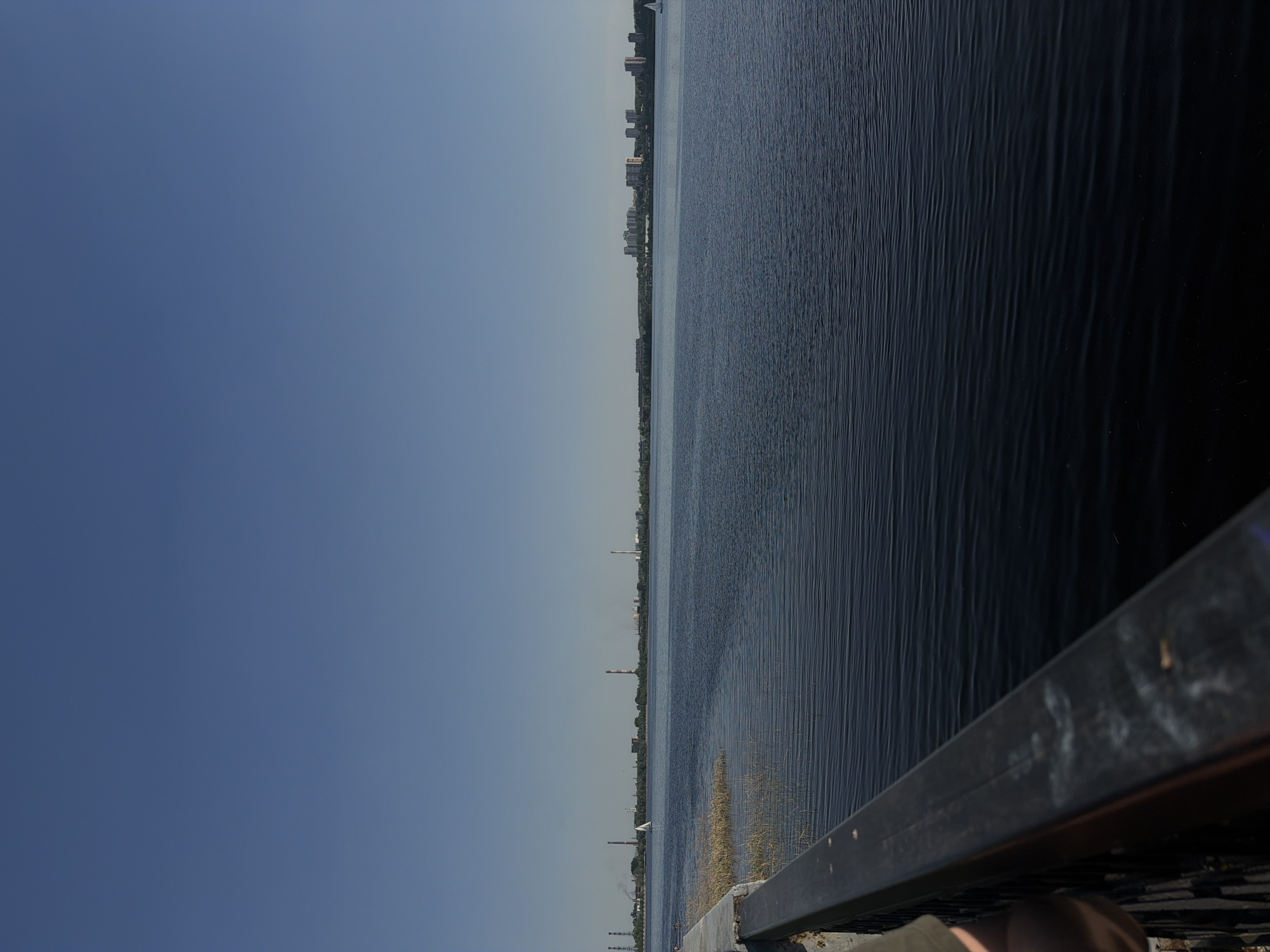 The image depicts a waterfront scene in Izhevsk, Russia. The left side of the frame is dominated by a blue sky. To the right of this is a body of water, likely the Izhevsk reservoir, with visible ripples on the surface. A cityscape, possibly of Izhevsk, is visible in the distance along the horizon. In the bottom right corner is a section of a metal railing, suggesting the image was taken from a walkway or bridge. A person is present on the bottom of the image. Several industrial structures, including chimneys, are visible along the shoreline. There is no interaction between people or activities are visible, and the environment appears to be a clear day.
