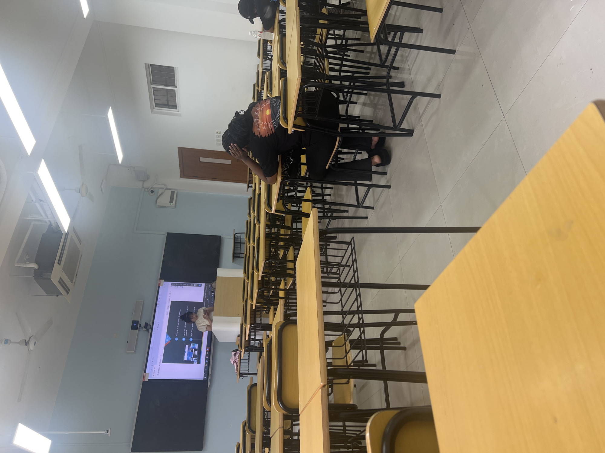 The image depicts a classroom setting with a presentation projected onto a large screen at the front. A person is standing at a lectern, seemingly addressing the class. Several students are seated at desks, with one student in the foreground appearing to be in distress or deep thought, resting their head in their hands. The classroom is equipped with typical school furniture, including desks, chairs, and overhead lighting. The overall atmosphere suggests a lecture or class session is in progress.