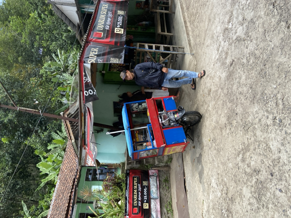 The scene is a street in Cipatat, Indonesia, located at latitude -6.796 and longitude 107.442. There are two people, both male, standing in front of a small shop. One man is wearing a black jacket and blue jeans, and the other is wearing a blue shirt and a black vest. Both men are standing in front of a small, red, blue, and yellow food cart with an attached motorcycle. The cart is being used to sell food, possibly "Diarum Super" as indicated on the signage. There are power lines running over the shop. The shop has a sign that reads "Diarum Super," and there are other signs with various advertisements. There are small tables and chairs placed outside the shop. Green trees and foliage are visible in the background. The ground is made of gray concrete.