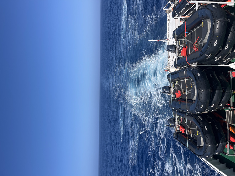 From a ship's perspective, the vast, deep blue ocean stretches to the horizon under a clear, cloudless sky. Turbulent white water churns in the ship's wake. Stacked black inflatable rafts are secured on the ship's deck, displaying the "Oceanwide Expeditions" logo. The rafts have red gas canisters, adding pops of color against the dark rubber.