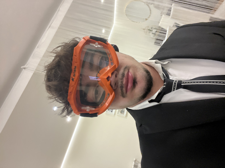 A man in a black suit and a black and white striped tie wears bright orange goggles. The goggles feature the brand name "Ariete" on the upper left corner of the frame. His brown hair is tousled. The background features bright white walls and decor, including a circular mirror and string lighting. The picture is taken at an angle, giving a skewed perspective.