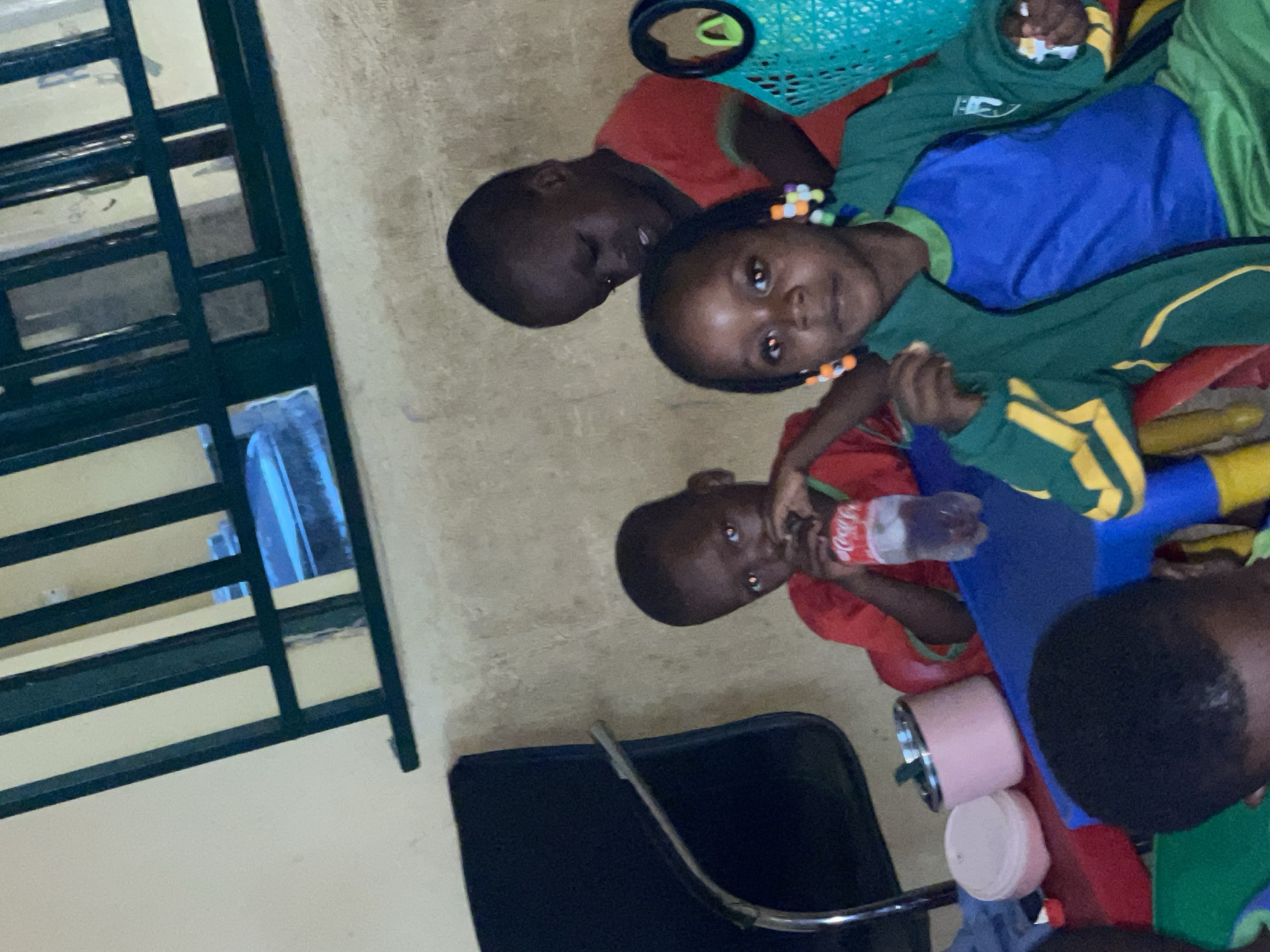 The image captures a group of at least four young children gathered indoors in what appears to be a classroom or playroom in Akwanga, Nigeria. The room has light-colored walls and a light-toned floor.
In the foreground, a girl wearing a green and blue uniform with colorful beads woven into her hair looks directly at the camera. Another child in a red shirt, positioned behind her, also looks towards the camera. A third child, also in a red shirt, is holding a clear plastic bottle with a red label displaying "Coca-Cola" and is drinking from it. Part of a fourth child's head, dressed in green, is visible in the lower right corner.
Various objects are present in the scene, including a prominent blue and red plastic play structure or toy in the center-right. A light blue/teal woven basket is visible in the upper part of the frame. Two pink cylindrical containers are placed on a surface near the children. A dark-colored chair with a metal frame is situated in the lower left corner. On the far left, a window with dark green metal security bars is visible.
The children appear to be engaged in a casual activity, possibly during playtime or a break, with several of them looking towards the camera with neutral to curious expressions. The indoor setting and lighting do not provide specific cues about the time of day.