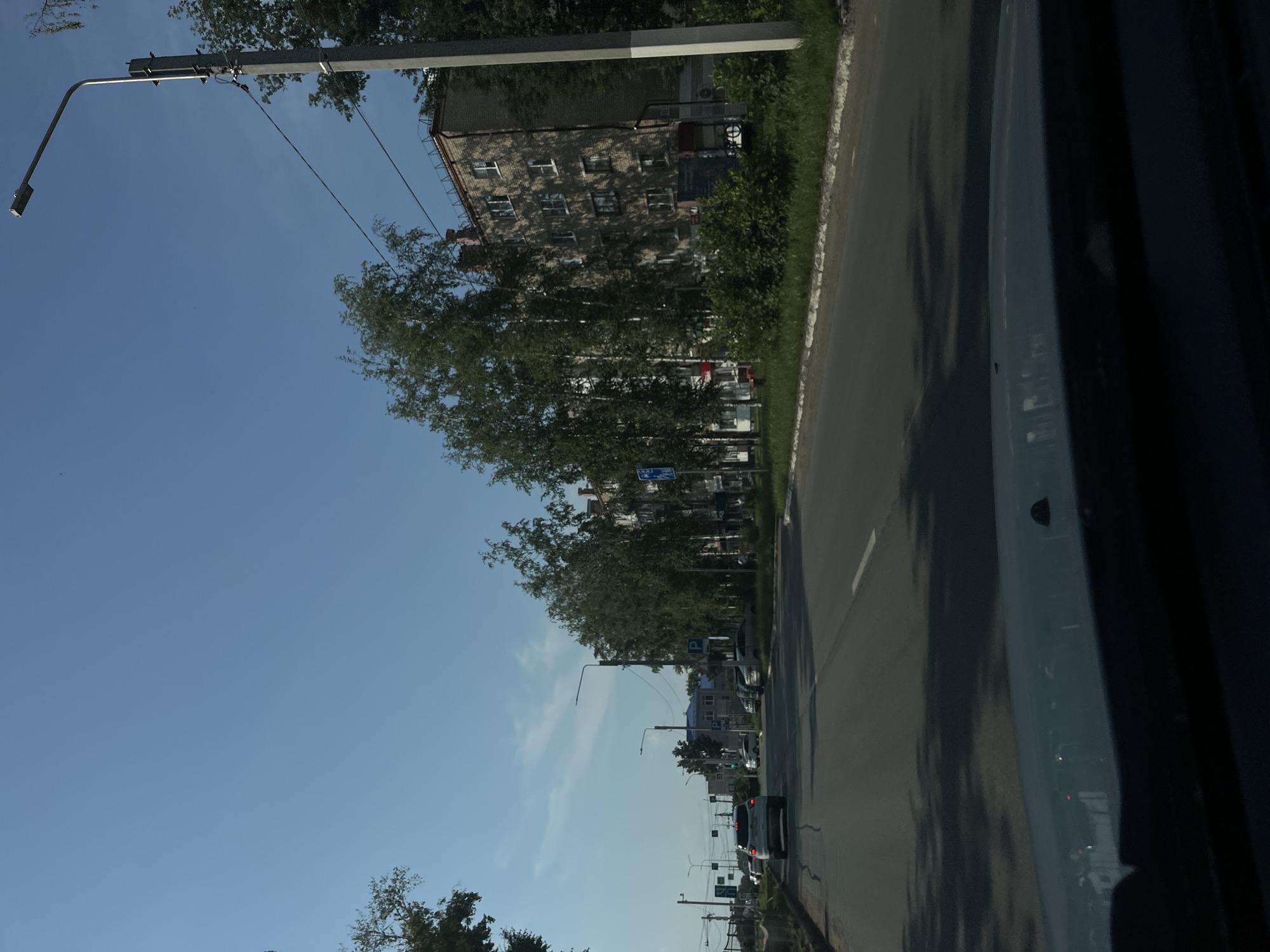 The image depicts a street scene in the city of Chaikovsky, Russia, under a clear blue sky. A paved road with lane markings runs through the center of the frame, with vehicles driving on it. On the right side of the road, a multi-story brick apartment building is visible, with several windows and what appears to be a pitched roof. Trees with light-colored bark and green leaves line the street in front of the building. A tall, modern street lamp stands prominently on the right side of the road, casting a shadow. On the left side of the road, traffic lights, signs, and more trees are present. The foreground shows the interior of a vehicle, with its dashboard and windshield visible, suggesting the image was taken from the driver's perspective. The overall impression is of a typical urban or suburban street on a sunny day.