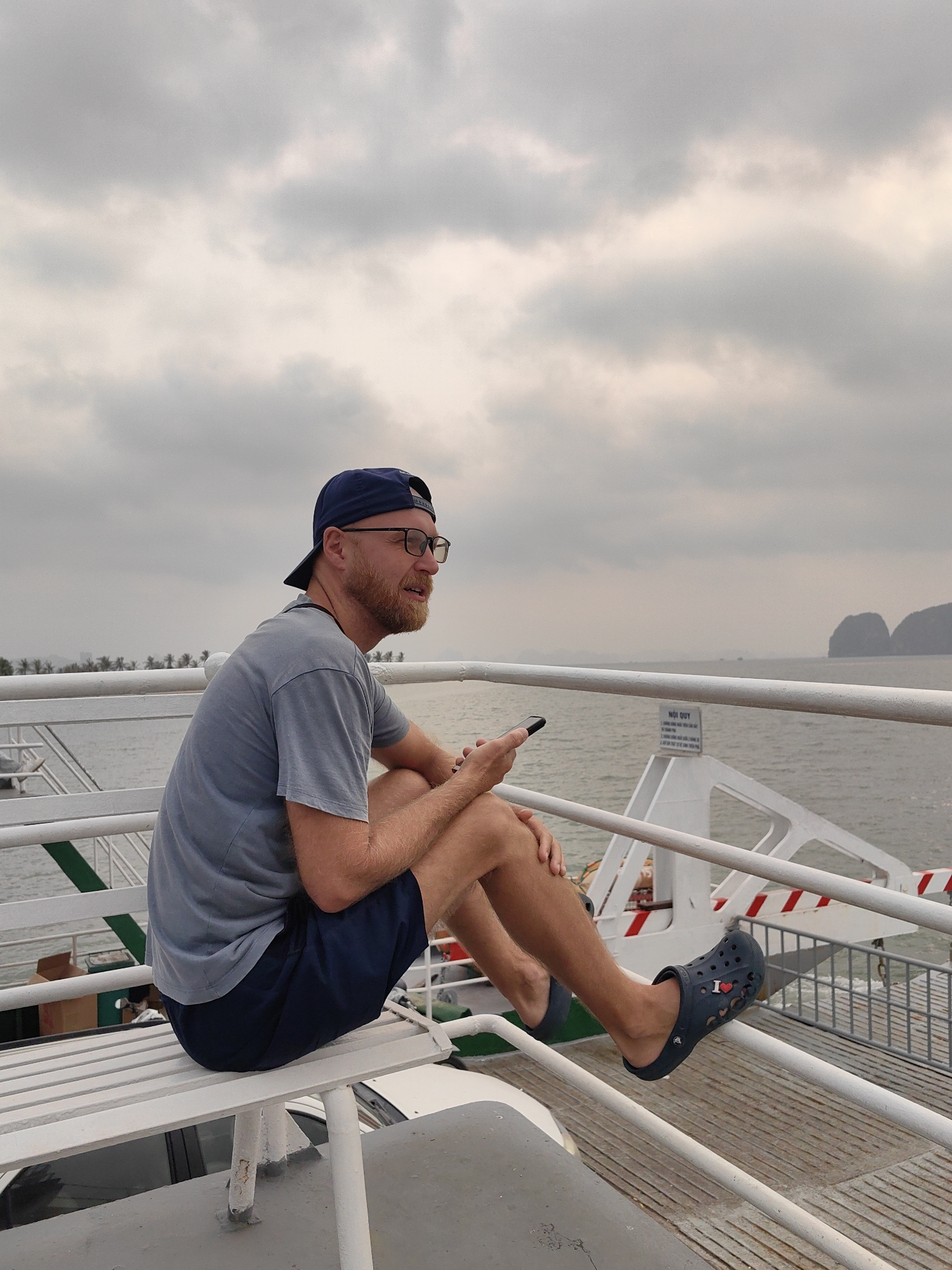 A full shot depicts a man with a beard wearing a navy blue baseball cap and gray shirt seated on a white bench on the deck of a ferry. He also has on navy blue shorts and dark blue Crocs with "I LOVE" logos. He is holding a mobile phone in his hand and gazing off in the distance. The ferry railing is in front of him. The background reveals an overcast sky and ocean view with distant land masses. The photograph was taken in the city of Tuan Chau Ward, Vietnam.