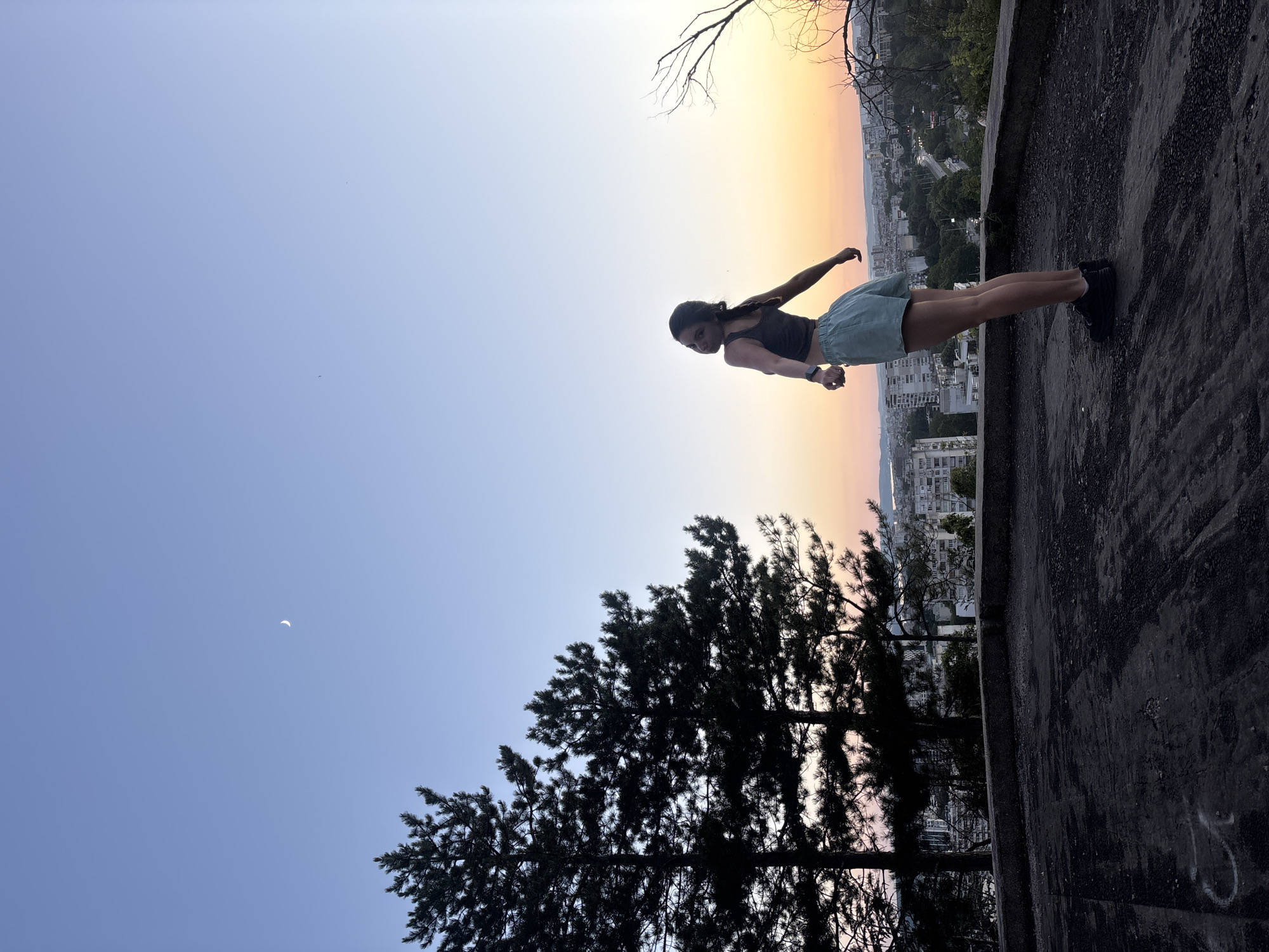 The image depicts a young woman standing on a dark, weathered concrete surface, possibly a rooftop or an elevated platform, overlooking a city at dusk. She is wearing a dark tank top and light blue shorts, with dark sneakers. Her body is turned away from the viewer, with her head looking back over her shoulder towards the camera. Her arms are extended outwards, and her posture suggests a pose of freedom or contemplation.
Behind her, the sky transitions from a clear blue at the top to a warm gradient of orange and pink near the horizon, characteristic of a sunset. A crescent moon is visible in the upper left quadrant of the sky. Silhouetted against the vibrant sky are several tall pine trees on the left side of the frame.
The city spread out below is characterized by a dense collection of buildings, some appearing to be apartment blocks, with visible roads and greenery interspersed. The urban landscape suggests Varna, Bulgaria, given the typical architecture and topography of the area. The overall atmosphere is one of calm and serenity, with the natural beauty of the sunset contrasting with the urban environment. The foreground concrete surface has some visible marks and debris.