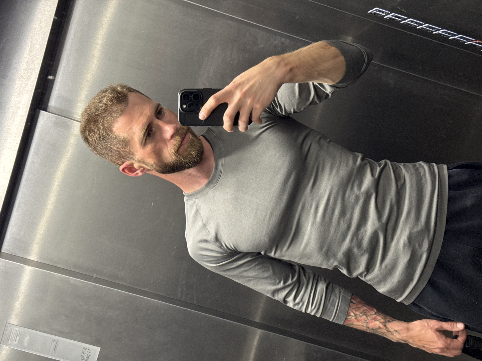 A fair-skinned male with short brown hair and a beard is taking a selfie in a modern elevator. He is wearing a gray long-sleeved shirt and dark pants. He is holding a black smartphone in his right hand. His left forearm shows a tattoo. The elevator has metallic walls with horizontal detailing at the top and bottom. There are also some stickers in the corner of one of the walls. The location cannot be verified in Budva, Montenegro, or anywhere else.