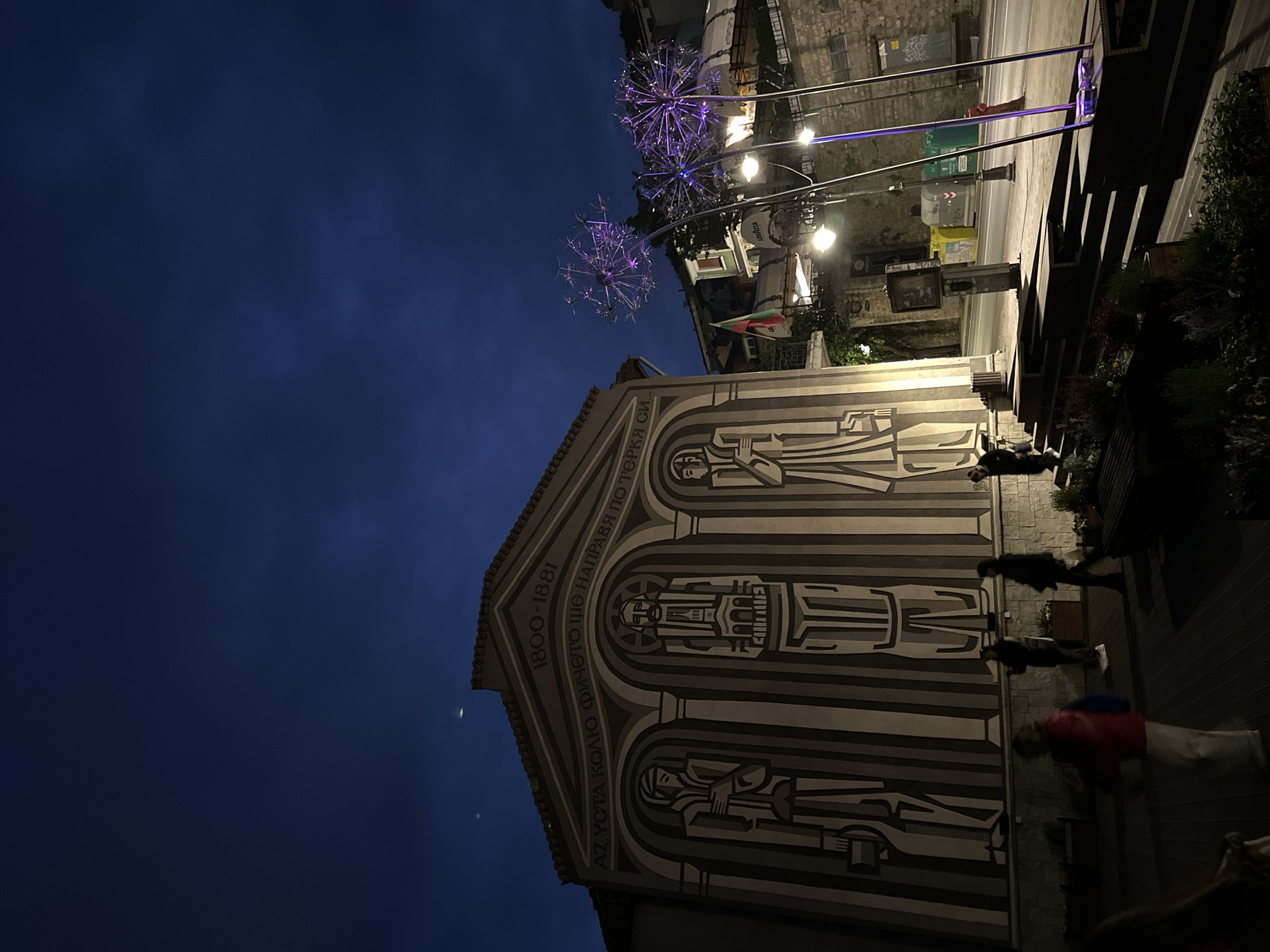 The image captures a nighttime scene in Veliko Tarnovo, Bulgaria, focusing on a building with a distinctive mural and street decorations. The building features a painted facade with stylized figures under arches, accompanied by the text "1800-1881 AZ, YCTA КОЛЮ Фичето, ше НАПРАВЯ ПО ТЕРКЯ СИ". The mural is vertically oriented in the image. A group of people is observed on the right side of the building, near what appears to be a staircase or wooden structure. Above the building, there are street lights with large, decorative spherical structures resembling dandelions or fireworks. In the background, there is a street with various street fixtures, including garbage cans, and additional buildings. A Bulgarian flag is visible near one of the decorative lights. The sky is a dark blue, suggesting a late evening or night setting.