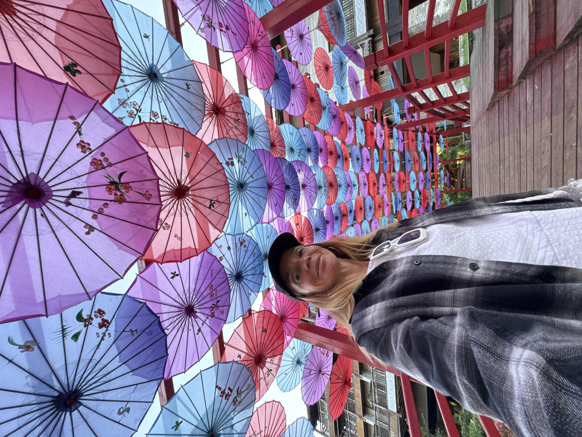 A photograph shows a woman wearing a black baseball cap, white shirt, and black and gray flannel shirt, standing beneath an array of colorful umbrellas suspended from a red framework. The umbrellas are various shades of blue, purple, and pink, some decorated with floral designs. The background reveals a partial view of a building with decorative wooden elements. The scene is located in Zhuoshui, China.