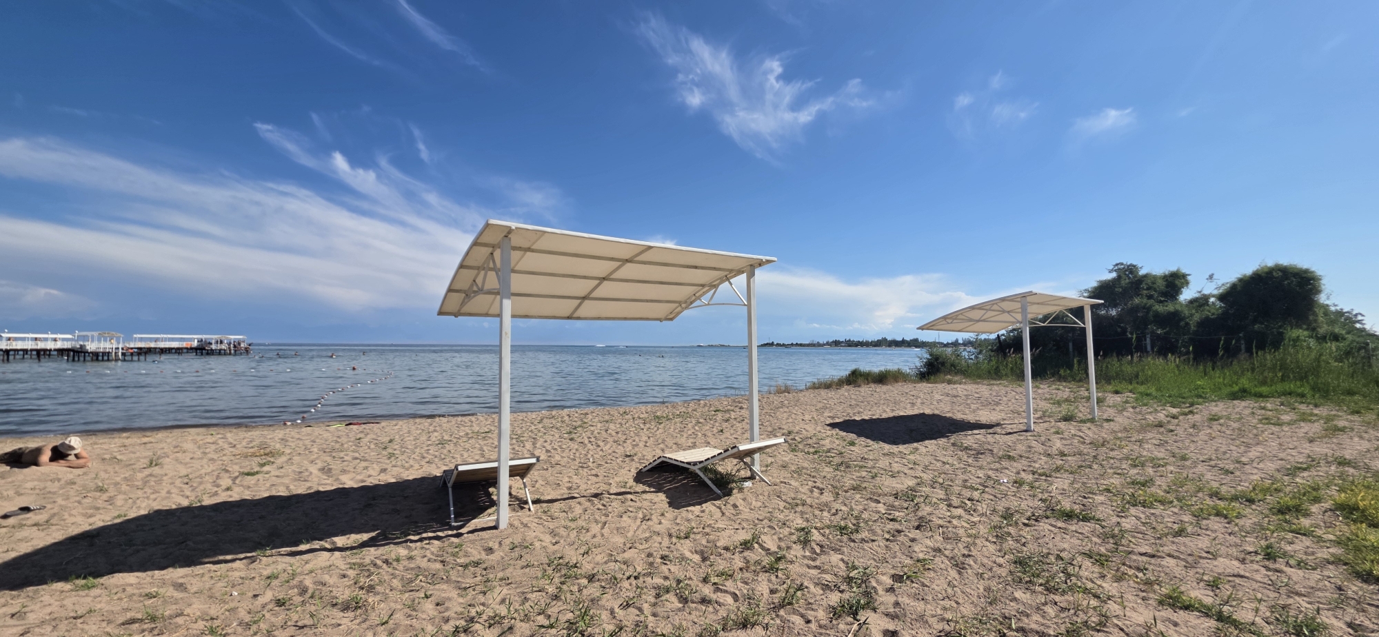 This is a wide-angle outdoor photograph taken on a sandy beach in the city of Choktal, Kyrgyzstan. The scene is dominated by a bright blue sky with wispy white clouds. In the foreground, a sandy beach is partially covered with sparse green grass. Two white, modern-style beach umbrellas with cream-colored canopies are prominent, each casting a shadow on the sand. Beneath the left umbrella is a collapsed white lounge chair, and beneath the right umbrella is another collapsed white lounge chair. A person in dark clothing and a light-colored hat is lying face down on the sand in the lower left corner of the image, with their legs extended towards the water.
The middle ground features the calm, blue water of a lake or sea, with a thin white buoy line demarcating an area. In the background, a long, white pier with multiple structures and people on it extends into the water. Beyond the pier and the water, the distant shoreline is lined with trees and vegetation, and a hazy mountain range is visible on the horizon. The overall impression is one of a serene and sunny day at a lakeside resort.