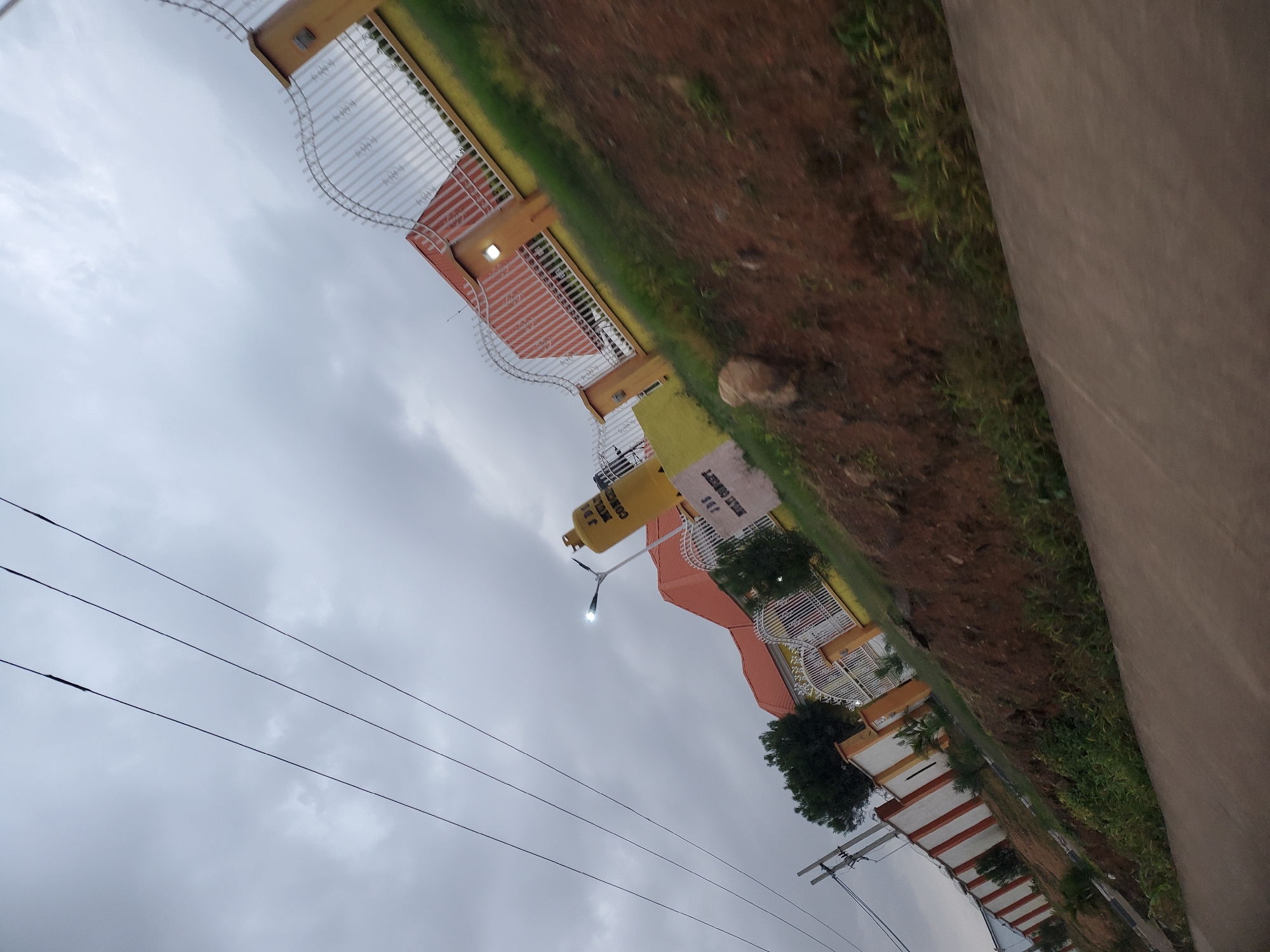 The image depicts the exterior of JJS MULTI CONCEPT in Gura Top, Nigeria. The scene is captured from a low-angle, tilted perspective, likely from a moving vehicle, with overhead power lines prominently crossing the sky. The cloudy sky dominates the upper portion of the frame.
The main subjects are the buildings and fences belonging to JJS MULTI CONCEPT. The architecture features a combination of orange and yellow structures, with white and orange striped walls. A large, yellow cylindrical object, possibly a billboard or a part of a sign, stands upright and displays text, partially visible as "JJS MULTI CONCEPT". A white, ornate gate with a pointed metal design is also visible. The property is bordered by a fence that transitions from white to yellow with decorative metal elements.
The foreground shows a dirt embankment covered with sparse green vegetation and a few rocks. A paved surface, likely a road or sidewalk, runs along the bottom of the frame. In the background, beyond the main building, is a densely vegetated area, with trees visible. A street light is illuminated and positioned above the entrance area. The overall environment suggests a semi-urban or developing area, with construction and power infrastructure present. There are no people visible in the image.
