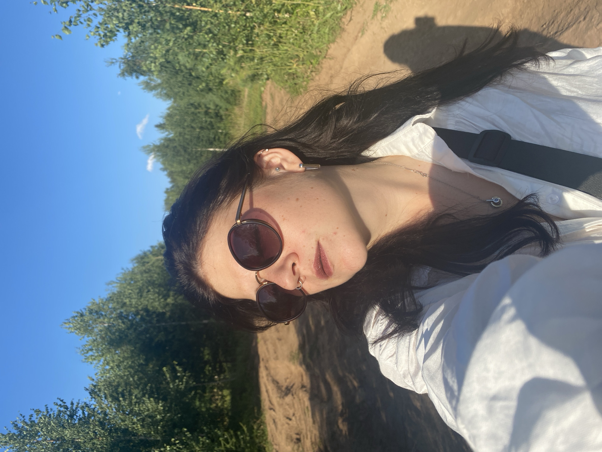 The media file is a portrait of a young woman in an outdoor setting. She has long, dark hair and is wearing sunglasses and a white shirt. The background consists of trees and a clear blue sky, suggesting a sunny day. The image is oriented sideways, with the woman's head towards the bottom right.
The subject appears to be taking a selfie, as indicated by the angle of the shot. The lighting suggests it is daytime, likely late afternoon or early evening due to the strong shadows. The environment seems to be a rural or natural area, possibly a park or a path lined with trees, consistent with the provided location context of Kostovaty, Russia.
Notable details include the woman's facial features, her earrings, and a necklace. The white shirt she is wearing is loosely buttoned and has shadows cast on it from the sunlight. The overall mood conveyed is casual and relaxed, perhaps indicating a moment of leisure or enjoyment of the outdoors. There is no visible text in the image.