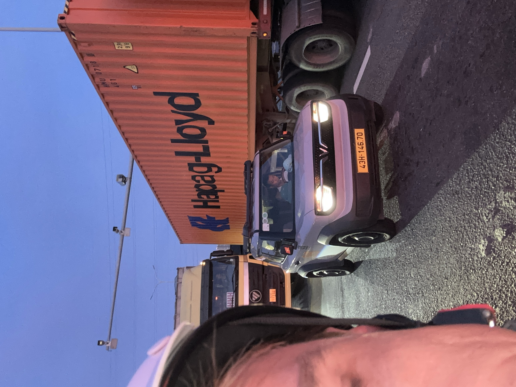 An exterior medium shot shows a traffic scene at dusk, possibly on a highway or major road in Da Nang, Vietnam. In the foreground, partially obscuring the left side of the frame, is the left shoulder and arm of a person wearing a helmet. The person is facing forward, and their head is slightly tilted.
In the middle ground, a light-colored electric vehicle, possibly a VinFast VF e34, is moving forward. It has its headlights on and a yellow license plate reading "43H-146.70". A male driver, wearing a white shirt and dark pants, is visible inside the vehicle.
To the right of the electric vehicle is a large, orange shipping container with the logo "Hapag-Lloyd" in dark blue lettering. The container is mounted on a truck trailer, and its rear wheels are visible. The container has shipping codes such as "HMU 07118 0" and "461" marked on it.
Behind the electric vehicle and to the left, a yellow truck is seen. Its front grille and headlights are visible.
Above the traffic, the sky is a clear, pale blue, transitioning into a darker hue at the top, indicating twilight. Two traffic cameras are mounted on a pole spanning across the road. The road surface is asphalt, with some white lane markings visible. The overall atmosphere is calm, with no immediate signs of distress or urgency.