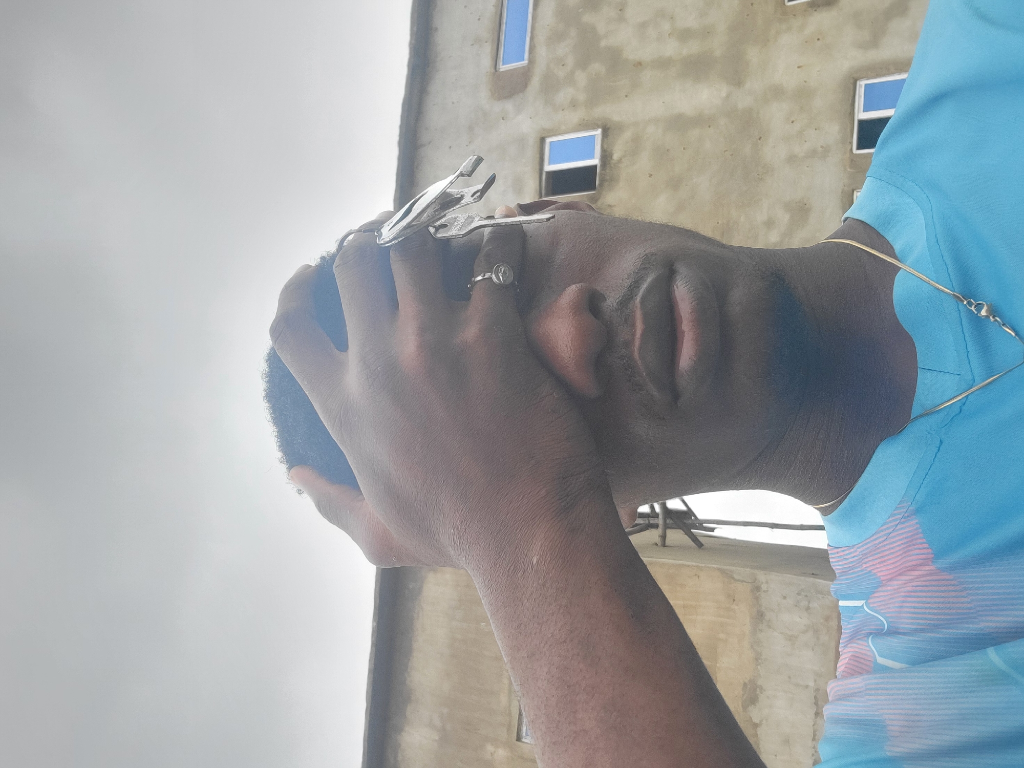 A dark-skinned individual is captured in a close-up, outdoor, selfie-style photograph. The person's right hand is raised, obscuring their right eye and forehead, and appears to be holding a set of keys. A silver ring is visible on the individual's left index finger. The individual is wearing a light blue t-shirt with a pink and white striped pattern. A thin gold chain is visible around their neck. The background consists of a dull, overcast sky and a textured, beige building with several rectangular blue-paned windows. Construction scaffolding is also visible against the building. The image is taken from a low angle, looking upwards at the individual. The overall scene suggests a casual, perhaps spontaneous, moment captured outdoors in an urban or semi-urban environment within Egbagu, Nigeria.