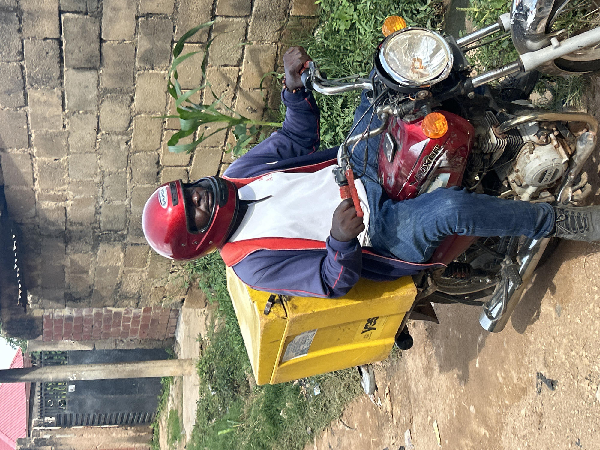 A person wearing a red helmet, blue and red jacket, and blue jeans rides a red motorcycle with a large yellow cargo box on the back. The motorcycle has a shiny chrome exhaust pipe and headlights. The person is looking forward, with their hands on the handlebars. The background consists of a brick wall, some greenery, and a dirt road. The overall scene appears to be in an outdoor urban or semi-urban environment, possibly in Jos, Nigeria.