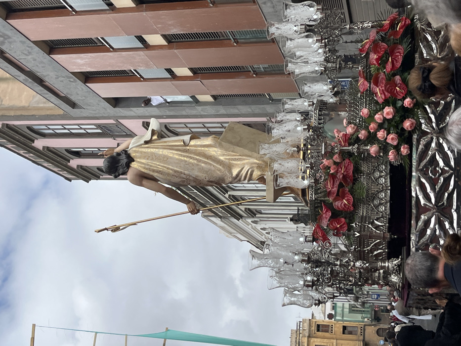 The image captures a religious procession in the city of Las Palmas de Gran Canaria, Spain. The procession is taking place on Calle de Triana, as indicated by the coordinates (latitude: 28.098, longitude: -15.416). The procession features a large, ornate float with a statue of Jesus Christ atop it. The statue is gold-colored and depicts Jesus with his arms raised in prayer. The float is decorated with numerous glass candle holders, red flowers, and white roses. The procession is moving down the street, and the image is taken from a perspective behind the float. Several people can be seen in the background, including a man in white clothing looking out of a window. A crowd of people, some obscured by the float, can be seen in the foreground watching the procession. The image is framed by the buildings on either side of the street. The buildings have multiple stories and feature balconies and windows. The sky is partly cloudy.