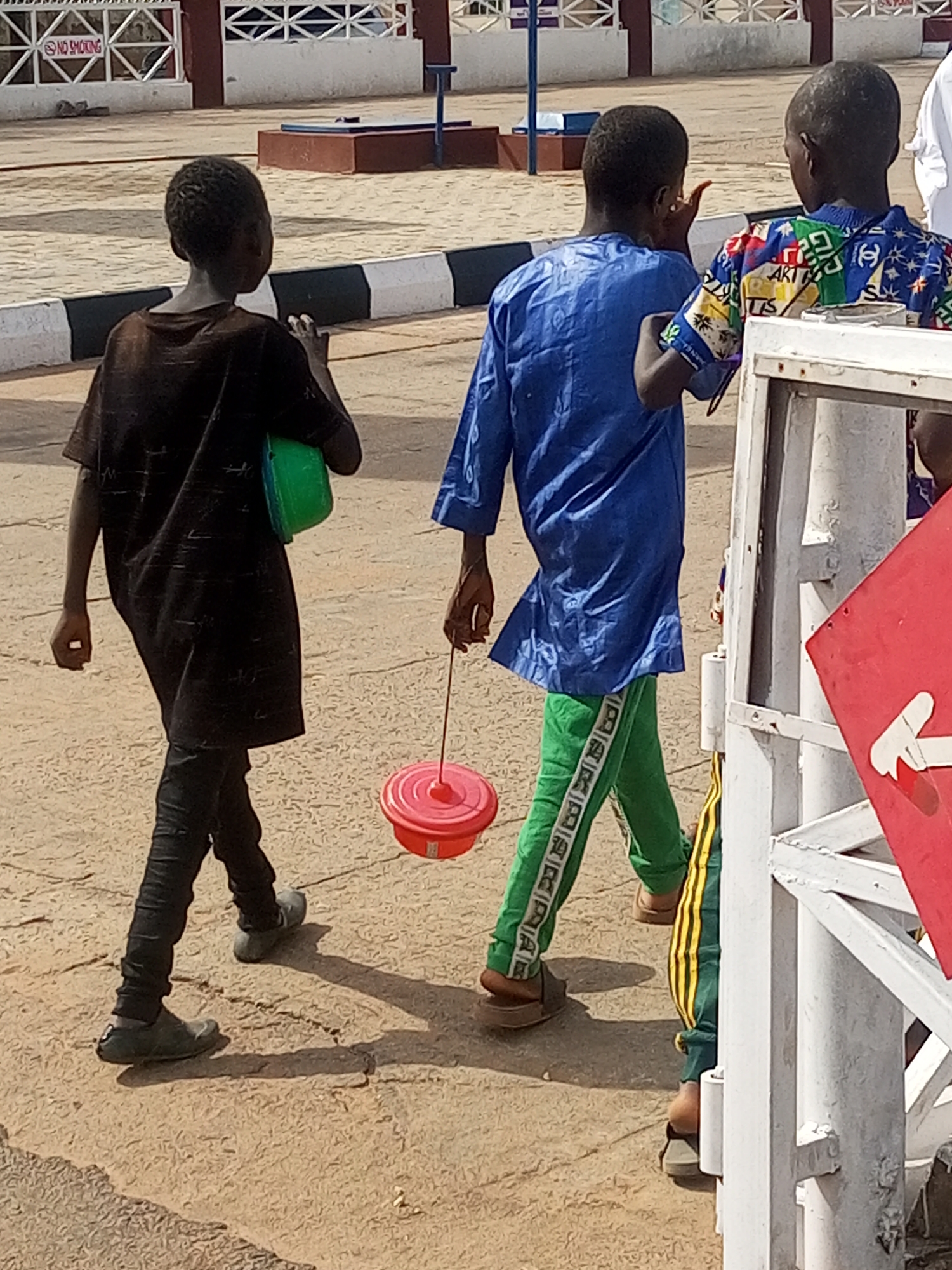 The image depicts a street scene in Gombe, Nigeria. Three young boys are walking on a paved surface with visible cracks and patches. The boy on the left is wearing a dark, loose-fitting tunic and black trousers, carrying a green container on his left side. The middle boy is dressed in a bright blue, long-sleeved traditional tunic and green trousers with white lettering down the sides. He is holding a red toy with a string in his right hand, dragging it behind him. The third boy, partially visible on the right, wears a colorful, patterned shirt. In the background, there is a white fence with the words "NO SMOKING" visible on a sign. The environment appears to be an outdoor, possibly public, area.