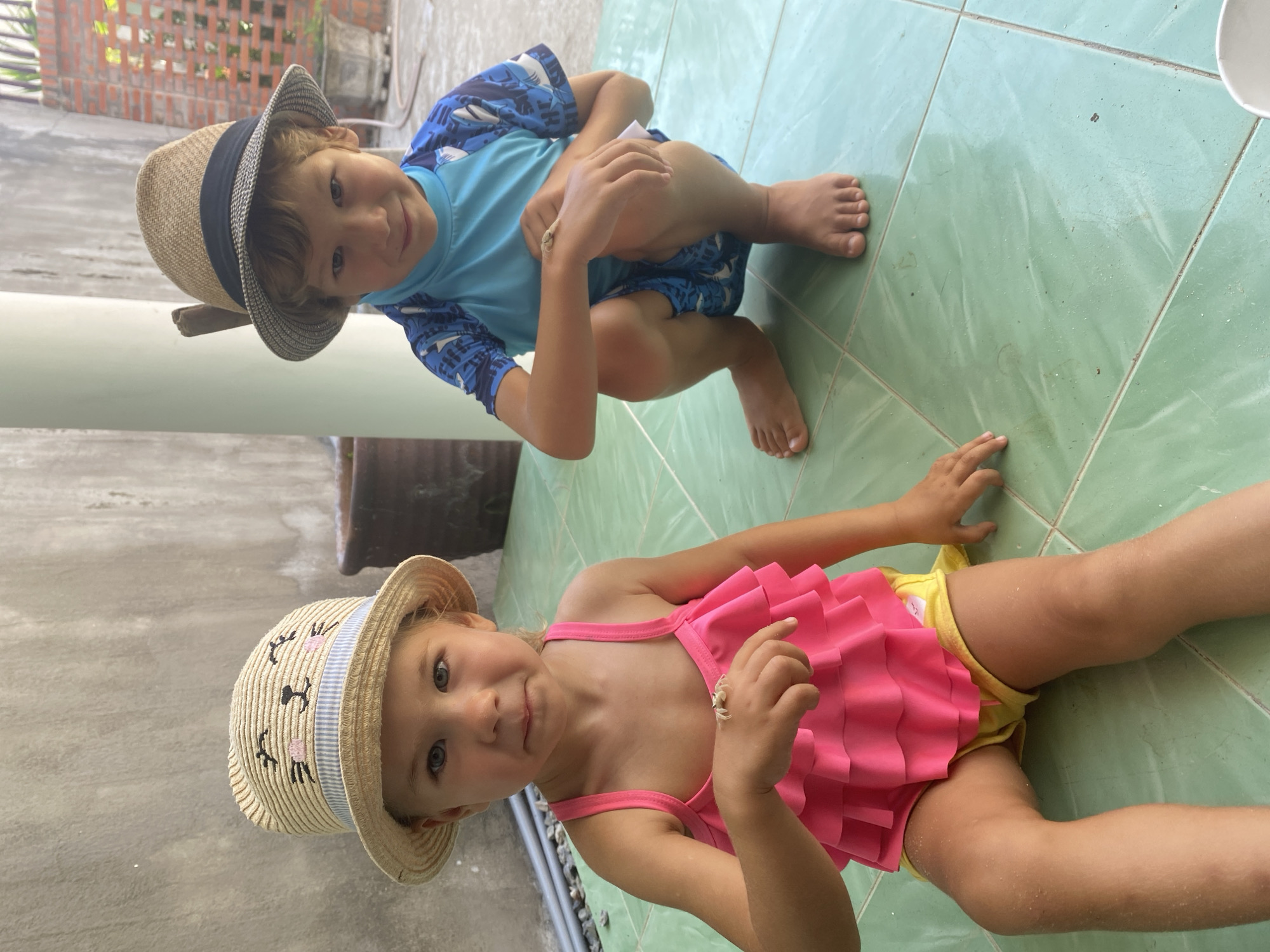 Two young children, a girl and a boy, are sitting on a light green tiled floor. The girl is on the left, wearing a pink swimsuit with ruffles and yellow shorts. She has a straw hat on her head decorated with a cat face. A small, light-colored crab is perched on her left hand. The boy is on the right, wearing a blue rash guard with a shark pattern and blue swim trunks. He has a brown and black straw hat on his head. A small, light-colored crab is on his right forearm. They appear to be outdoors, with a textured concrete wall and a brick wall visible in the background. A large brown pot is behind the girl, and some greenery and a white bucket are visible behind the boy. The overall scene suggests a warm, possibly coastal environment, consistent with Mũi Né, Vietnam, known for its beaches and resort areas.