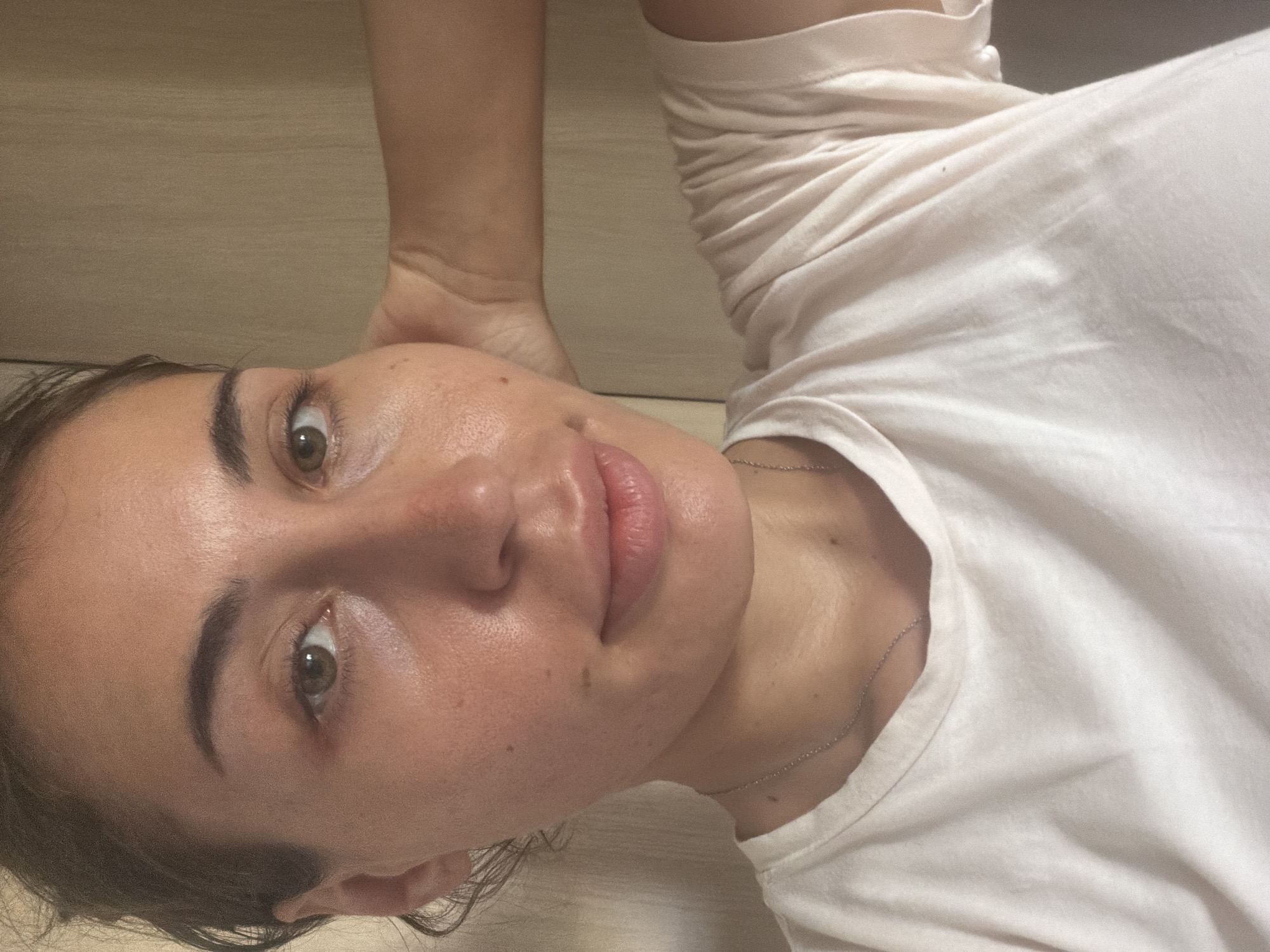 The image is a close-up selfie of a woman with light brown hair pulled back, wearing a light-colored t-shirt and a delicate silver necklace. She has brown eyes and is looking towards the camera. Her right hand is resting on her head, and her left arm is bent. The background appears to be an indoor setting with a wooden wall. The woman's skin appears to have a dewy glow. The scene is likely in a residential setting in Tersefanou, Cyprus.