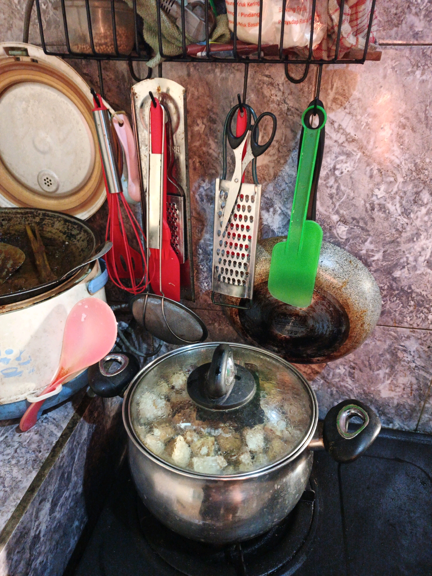A busy kitchen counter is seen in a medium shot, with various cooking utensils and pots. In the foreground, a silver pot with a glass lid is placed on a black stovetop. The pot contains food that appears to be simmering. To the left of the pot, a pink cooking spoon is partially visible, along with a white pot or container. Above these items, a set of kitchen tools hangs from hooks mounted on a textured wall. These tools include a red whisk, a red spatula, a grater, and a pair of scissors. A green spatula is also hanging, resting on a dirty, round pan. In the background, a shelf holds several jars and containers, one of which appears to contain small, brown items like seeds or grains. The wall has a marble-like pattern in shades of brown and gray. The overall scene suggests a working kitchen environment, possibly in a home or a small eatery. The specific location mentioned for Ketegan, Indonesia, is an external context not visually present within the image itself.