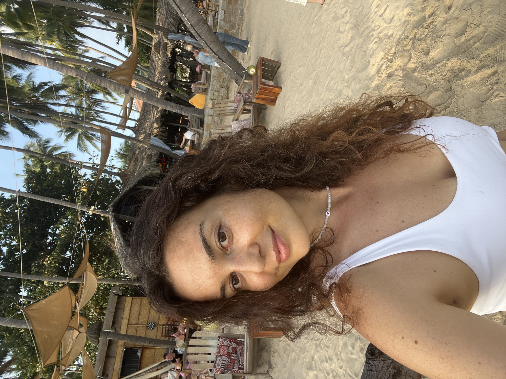 A young woman with long, curly brown hair and fair skin, adorned with a pearl necklace with a red heart pendant, takes a selfie on a sandy beach. She is wearing a white swimsuit. The background depicts a beachside establishment with thatched roofs, wooden chairs, and palm trees, suggesting a tropical resort or restaurant in Mũi Né, Vietnam. Other people can be seen in the background, some sitting at tables and others walking. The atmosphere appears relaxed and casual, typical of a beach vacation destination.