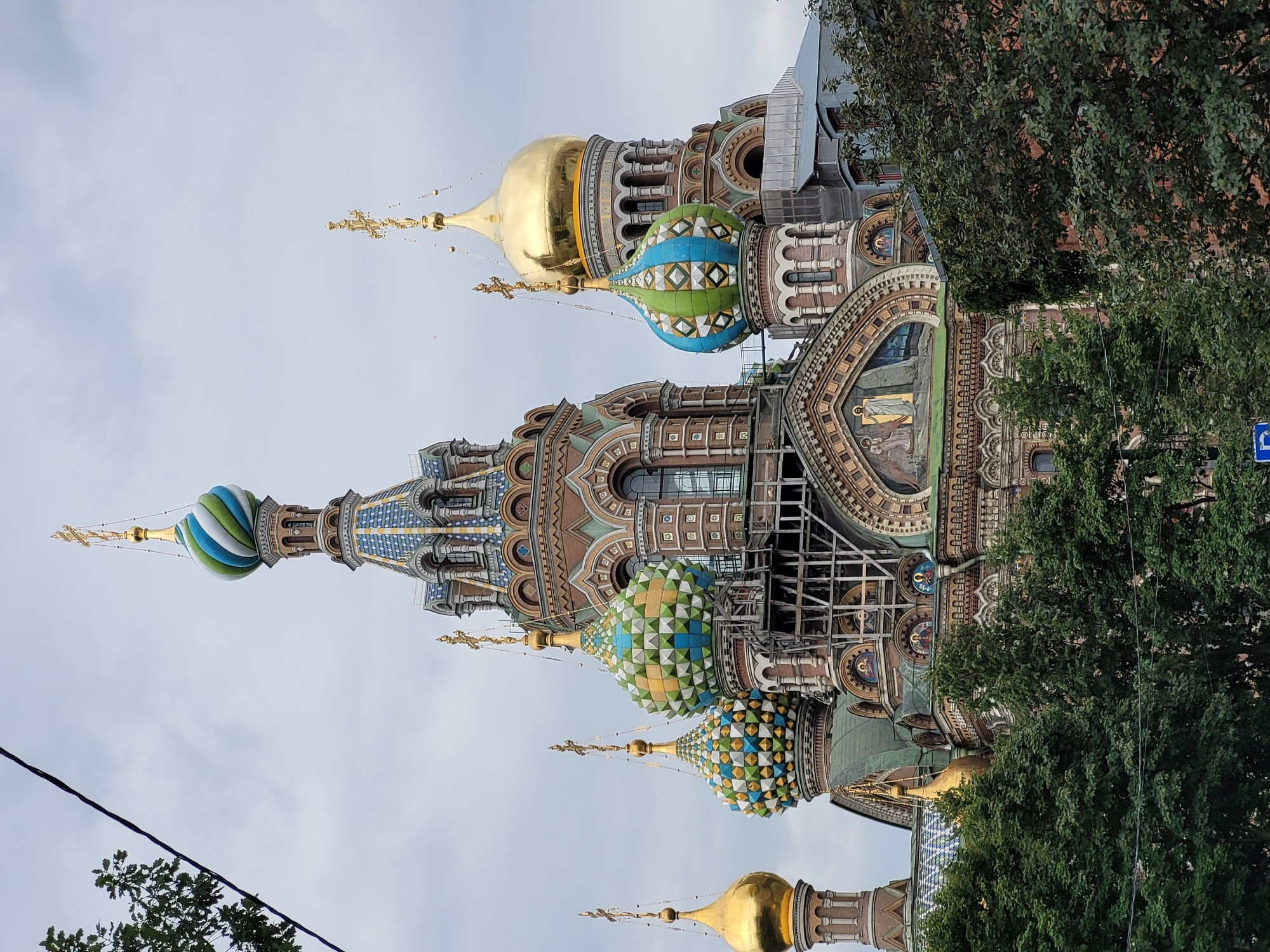 The image shows the Church of the Savior on Spilled Blood in Saint Petersburg, Russia. The ornate church features multiple onion domes, some gilded and others adorned with intricate blue, green, and gold patterns. The exterior of the church is richly decorated with colorful tiles and mosaics. Construction scaffolding is visible on one of the domes, suggesting ongoing restoration or maintenance. Lush green trees partially obscure the lower portion of the church. The sky is overcast, contributing to a muted lighting condition.
