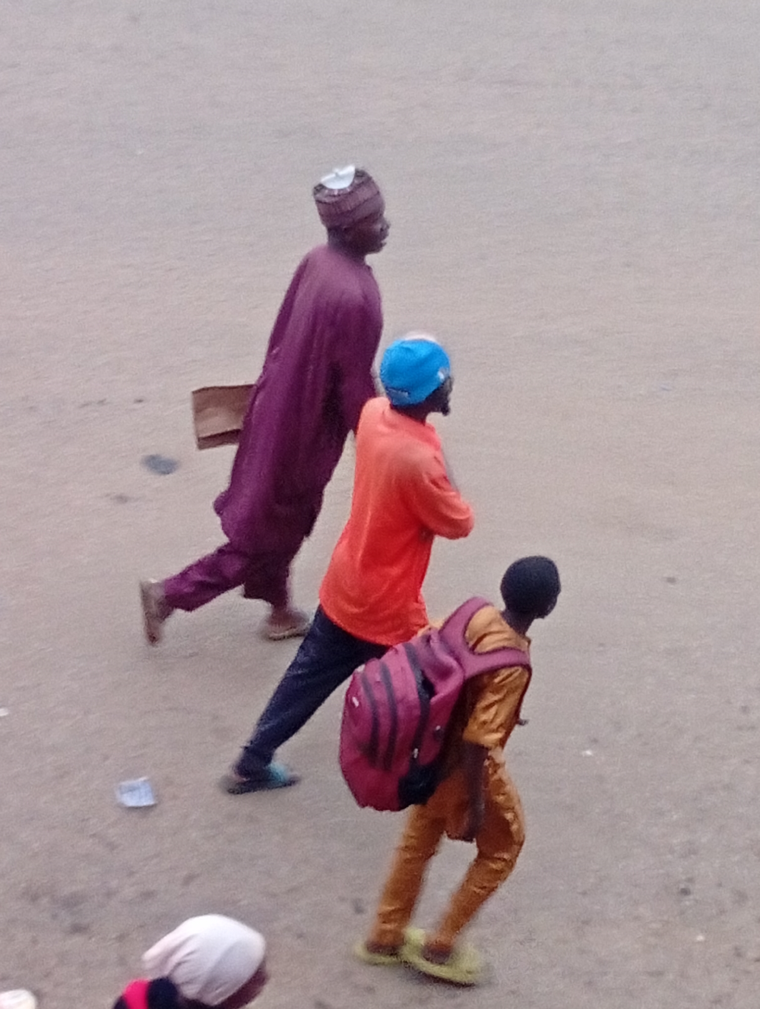 The image depicts a street scene in Bukuru, Nigeria, with three individuals walking from left to right. In the foreground, a person wearing a white head covering is partially visible, facing away from the camera. Behind them, a young person in a yellow shirt and brown trousers is walking, carrying a maroon backpack. Further back, an individual in an orange shirt and dark trousers is walking, wearing a bright blue helmet. The farthest person on the left is wearing a dark purple traditional garment and a patterned cap, carrying a brown paper bag. The ground appears to be a paved surface, possibly a road or walkway, with scattered debris. The lighting suggests it is daytime.