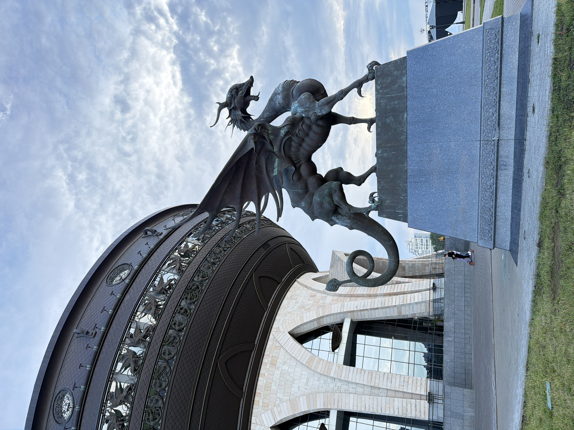 This media file showcases an impressive outdoor sculpture of a dragon, a prominent landmark in Kazan, Russia. The dragon, depicted in a dynamic pose as if poised to strike, is made of dark metal and features intricate detailing on its wings, scales, and sharp claws. It is mounted on a large, grey granite pedestal. The sculpture is situated in front of a modern building with a distinctive, curved, dark metal roof and a facade clad in light-colored stone. Large glass windows reflect the sky and surrounding environment. To the right of the sculpture, a paved walkway leads past a grassy area. A person is visible in the distance, riding a scooter on the walkway, adding a sense of scale to the scene. The sky is partially cloudy, suggesting it might be daytime with a mix of sun and clouds. The overall atmosphere is one of grandeur and architectural interest, highlighting the dragon sculpture as a central feature of the urban landscape.