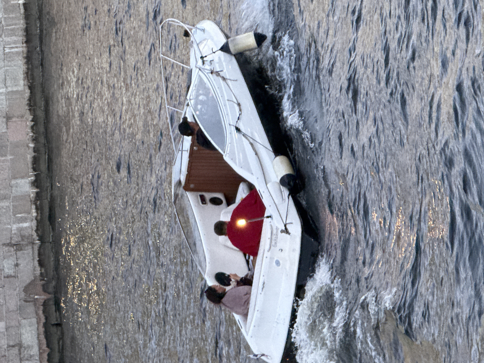 The image captures a white motorboat navigating a body of water, likely a river or canal, with a stone embankment visible in the background. The embankment, constructed of large, rectangular stone blocks, suggests an urban setting. The water is choppy, indicating the boat's movement, and reflects light, hinting at either sunlight or artificial illumination.
Inside the boat, three individuals are seated. On the left, a person wearing a light-colored wrap is looking downwards. Next to them, a person in a bright red garment is facing away from the camera, seated on the right side of the boat's rear. In the middle, another person, partially obscured, is seated behind the individual in red. The boat itself has a windshield, a railing, and two cylindrical buoys attached to its sides, one on the port side and one on the starboard side, both exhibiting black and yellow markings. A speaker unit is visible on the interior wall of the boat. The boat's name, "Sunshine," is inscribed on its hull. The scene appears to be set in Saint Petersburg, Russia, given the architectural style of the embankment and the common presence of such waterways within the city.