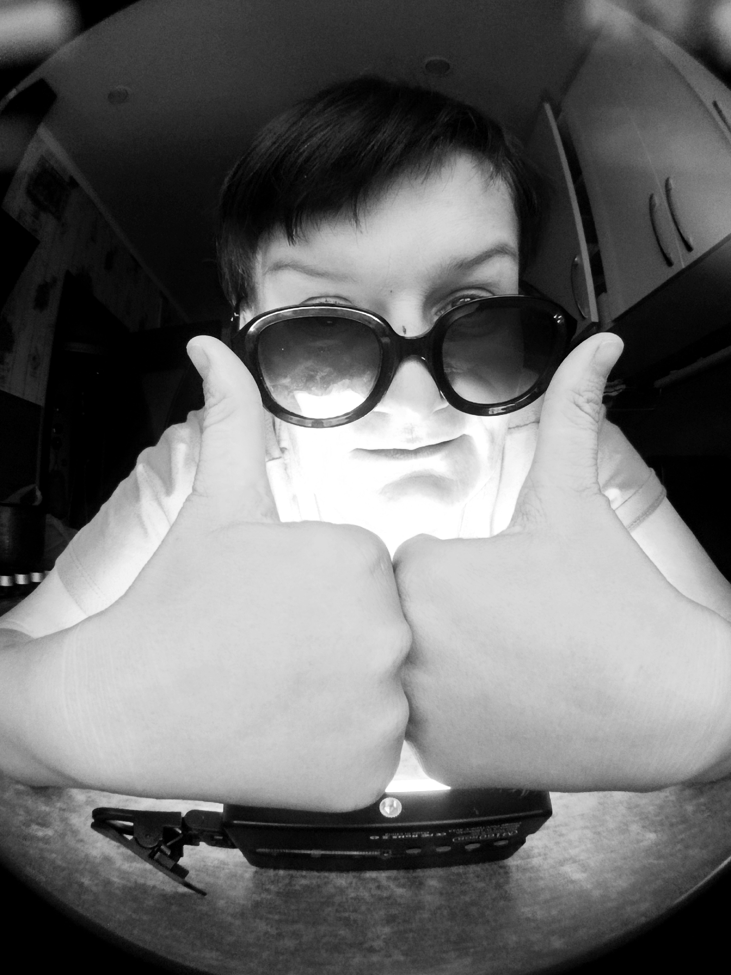 The image is a black and white, fisheye perspective selfie of a person wearing large sunglasses. The person is holding their hands up in front of their face, giving two thumbs up. The background is dimly lit and appears to be an indoor environment, possibly a room in a house. There are cabinets and a ceiling with lights visible. A black electronic device with a clip is visible on the surface below the hands. The image does not provide any information that would allow for the determination of its location in the city of Фоки, Russia.