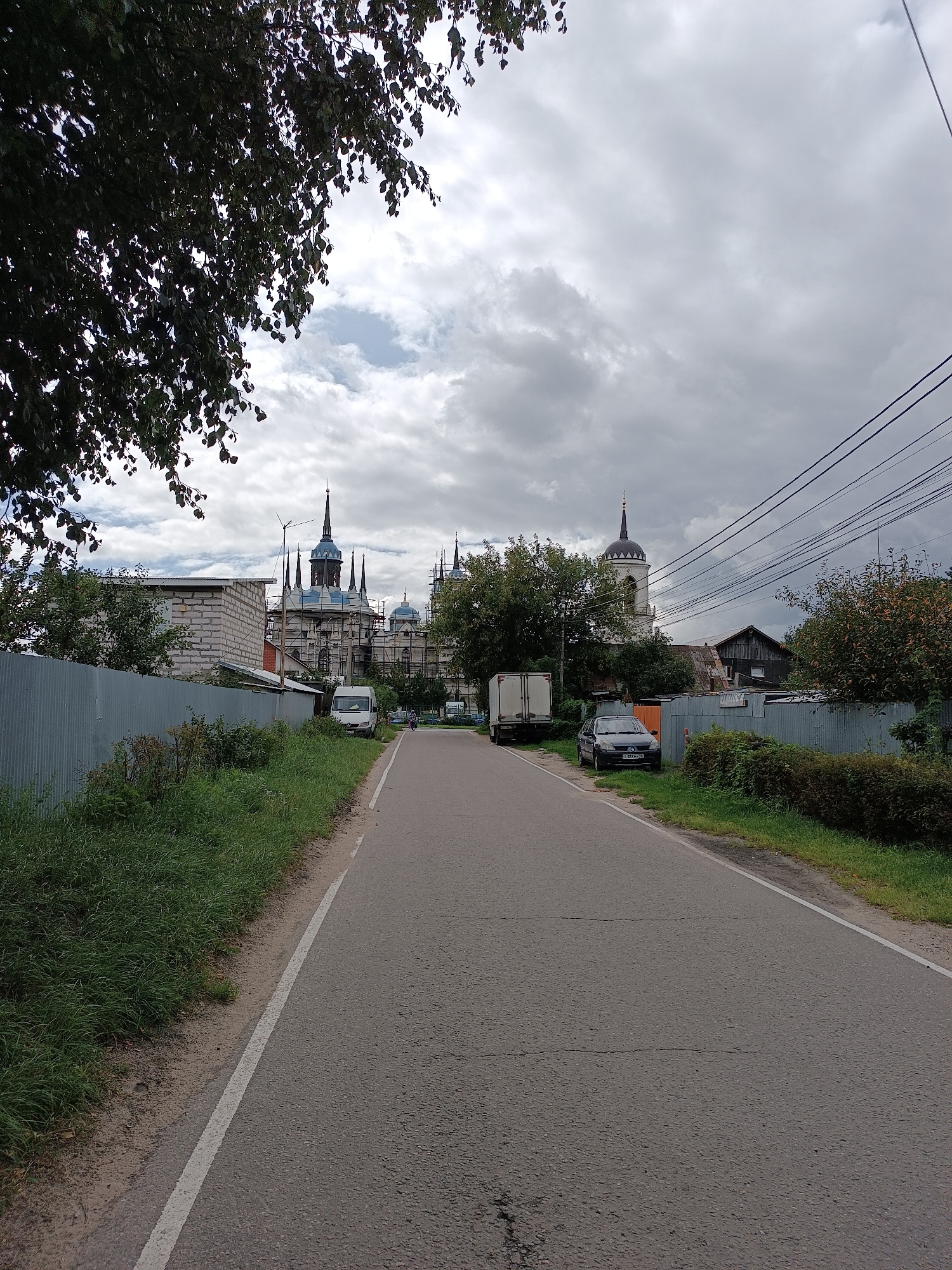 The image depicts a street scene in the village of Bykovo, Russia, under a cloudy sky. A paved road runs through the center of the frame, bordered by a grassy verge and a corrugated metal fence on the left, and more grass, hedges, and a fence on the right. In the distance, on the left side of the road, a large building with multiple spires and blue domes, characteristic of Russian church architecture, is under renovation with scaffolding. To the left of this building, a brick structure is visible. Further down the road, a white van is parked, and a person is seen riding a bicycle. On the right side of the road, a dark sedan is parked next to a fence, and a white truck with a cargo box is parked further ahead. A bell tower with a dark dome is also visible on the right, behind trees and houses. Electrical wires crisscross the sky in the upper right portion of the image. The overall atmosphere is subdued due to the overcast weather.