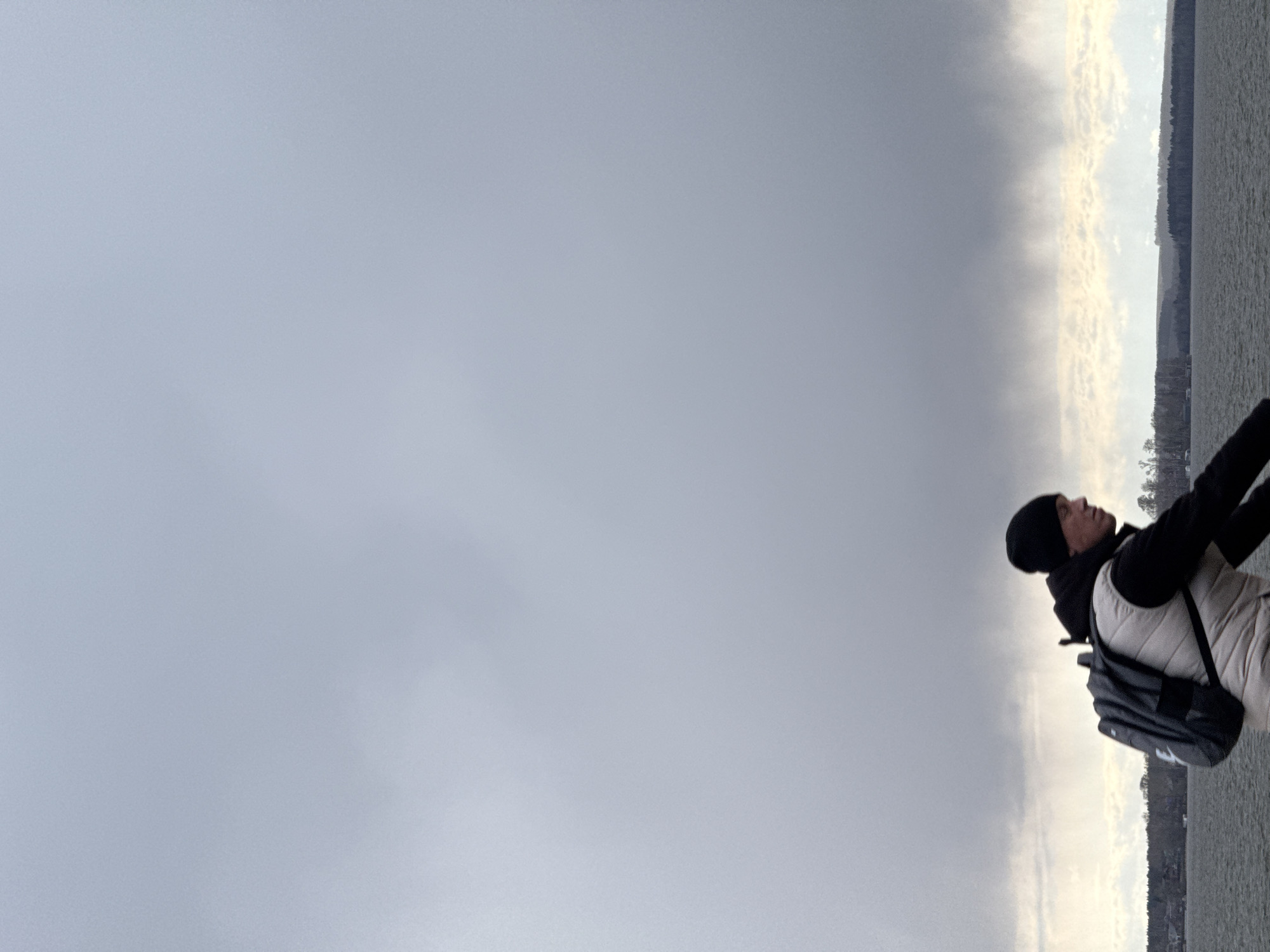 A person stands on the right side of the frame, gazing upwards and to the left. The individual is dressed in cool-weather attire, including a black beanie, a dark hooded top, a light-colored (possibly off-white or beige) puffy vest, and a dark grey backpack worn over the vest. Their arms are slightly extended, implying a posture of observation or interaction with the expansive environment.
The background features a broad body of water, likely a lake or reservoir, spanning the lower half of the image and appearing calm. Across the water, a dark, distant tree line marks the horizon. The sky dominates the upper and left portions of the frame, presenting a mostly overcast appearance. It transitions from a solid light grey on the far left to slightly brighter, more textured clouds closer to the horizon, suggesting a mix of cloud cover.
The scene conveys a cool, overcast daytime atmosphere. The clothing of the person suggests chilly temperatures. The setting is an outdoor landscape beside a large body of water, consistent with a location like Votkinsk Reservoir in Russia. No visible text is present in the image.