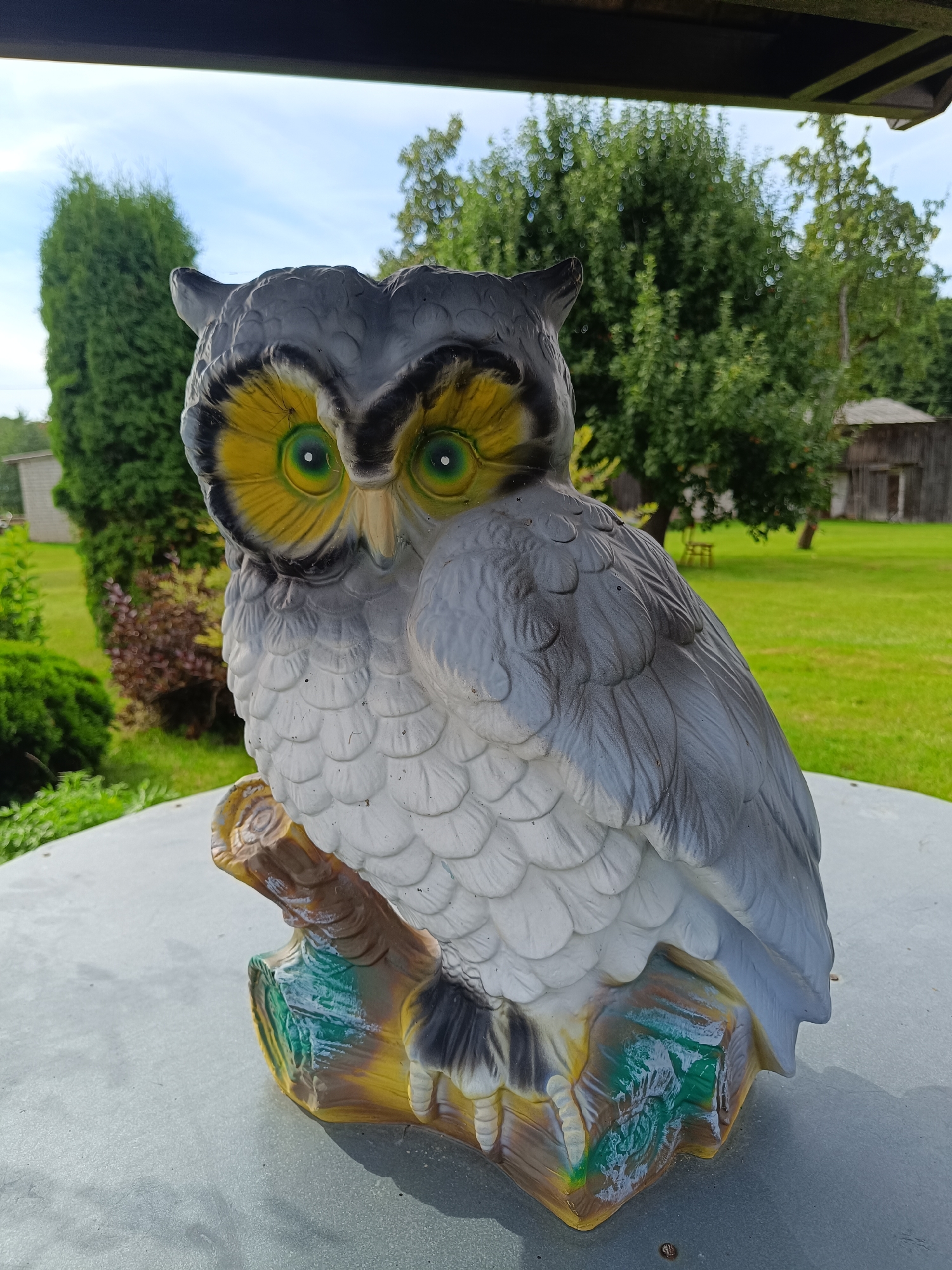 An owl statue is positioned on a round, light gray surface, likely a table, in an outdoor setting. The statue, primarily gray and white with vibrant yellow and green eyes, depicts an owl perched on a stylized branch with colorful accents of yellow, brown, and green. The background reveals a green lawn with scattered trees and bushes, and a wooden structure in the distance. The sky above is a clear, light blue with some white clouds. The location is identified as Tebras, Latvia. There are no people or animals actively engaged in any interactions within the frame.